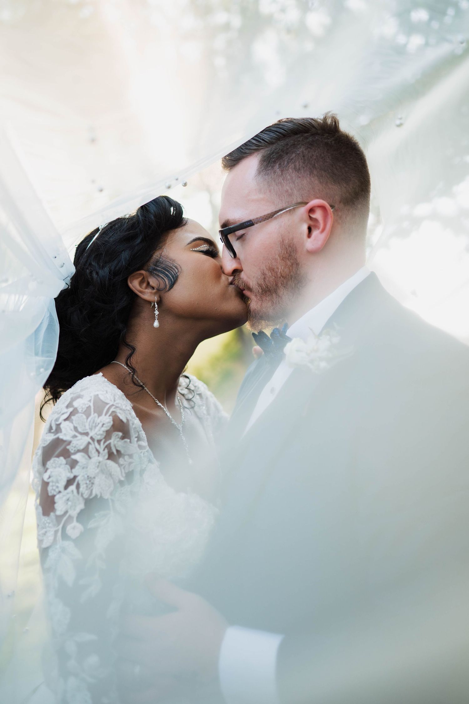 Bride and groom share a romantic kiss under a flowing white veil on their wedding day.