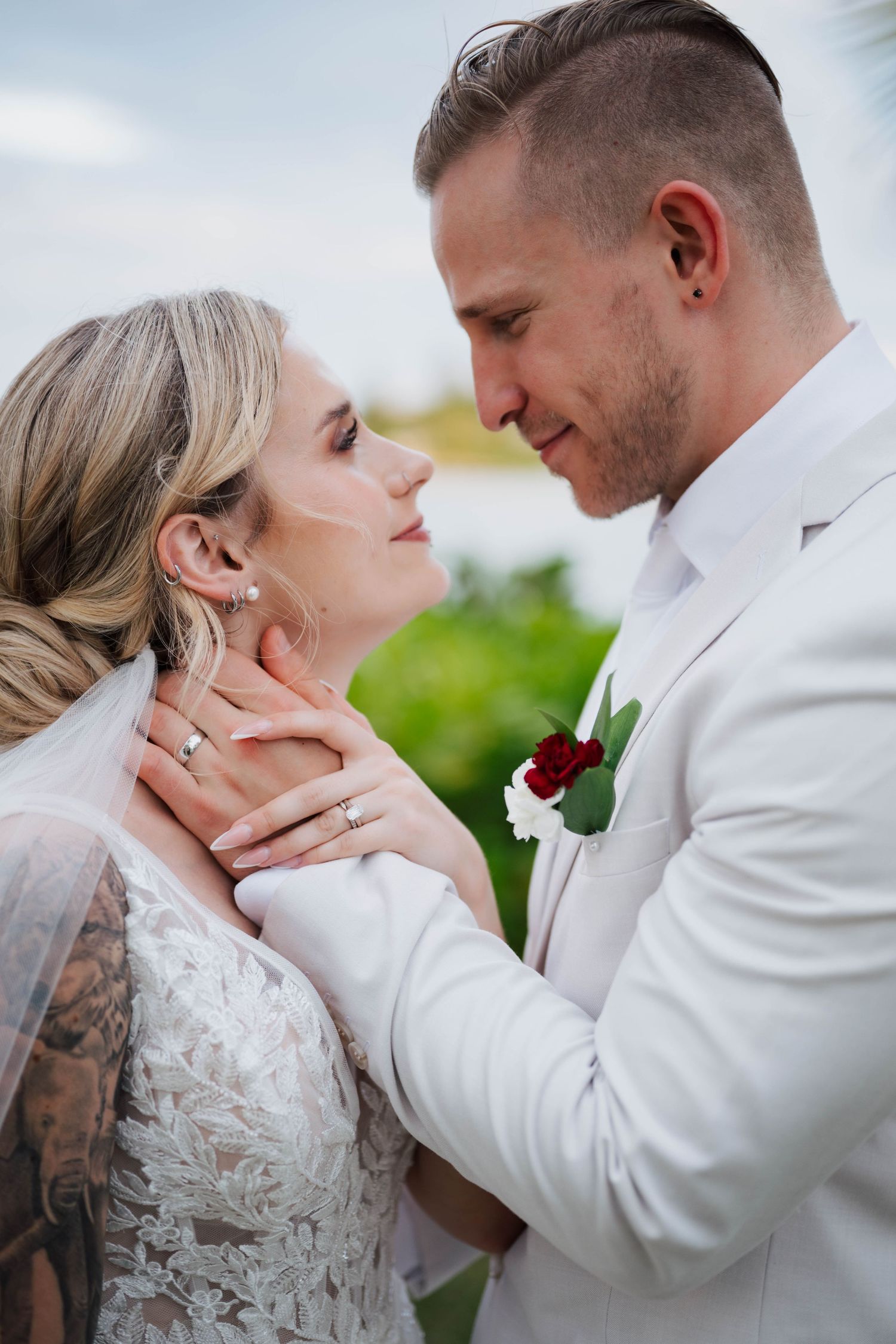 Bride and groom share a romantic moment, groom tenderly holding brides face, wearing white suit with red boutonniere.