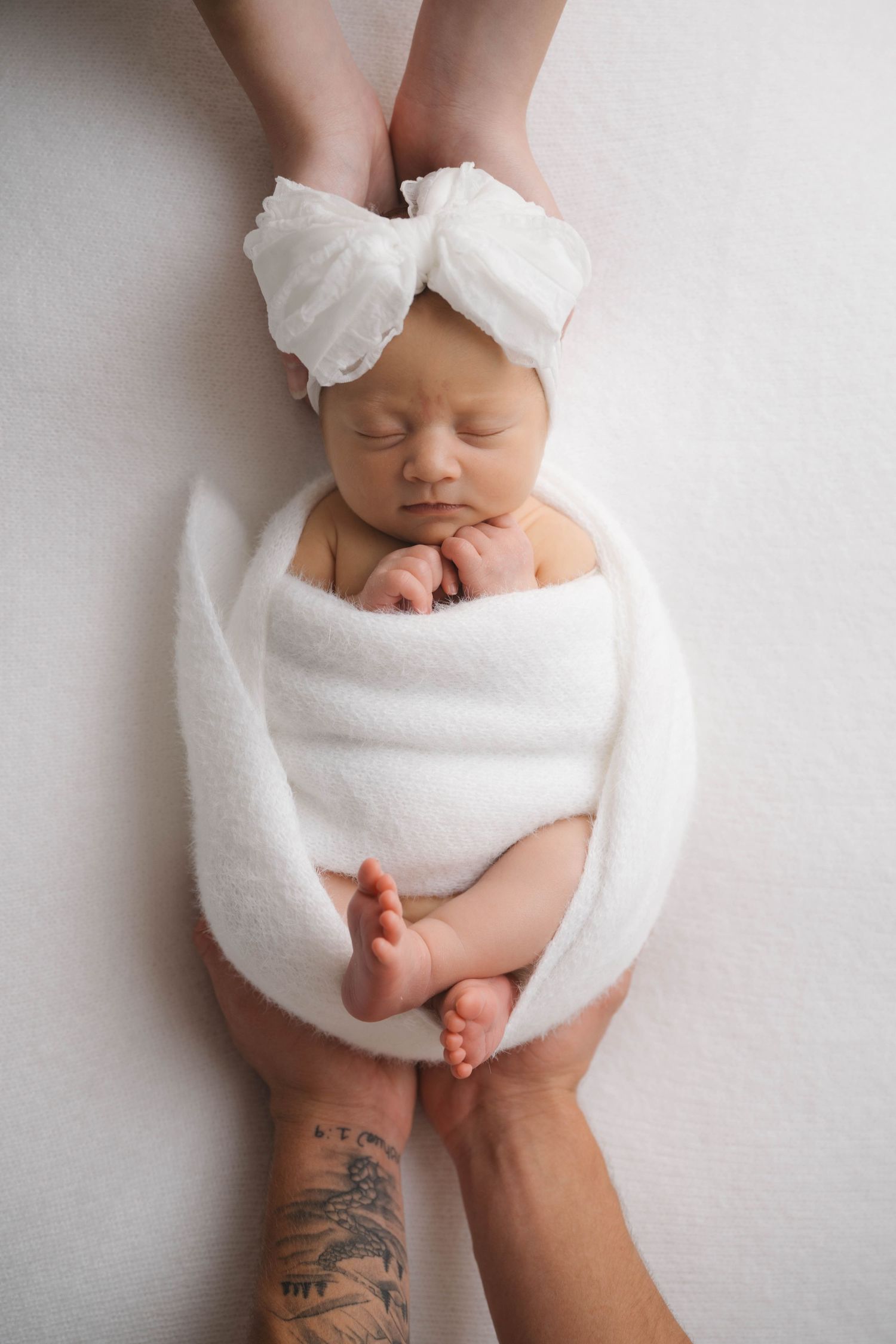 Newborn baby girl wrapped in white blanket with bow headband, cradled in parents hands on white background.