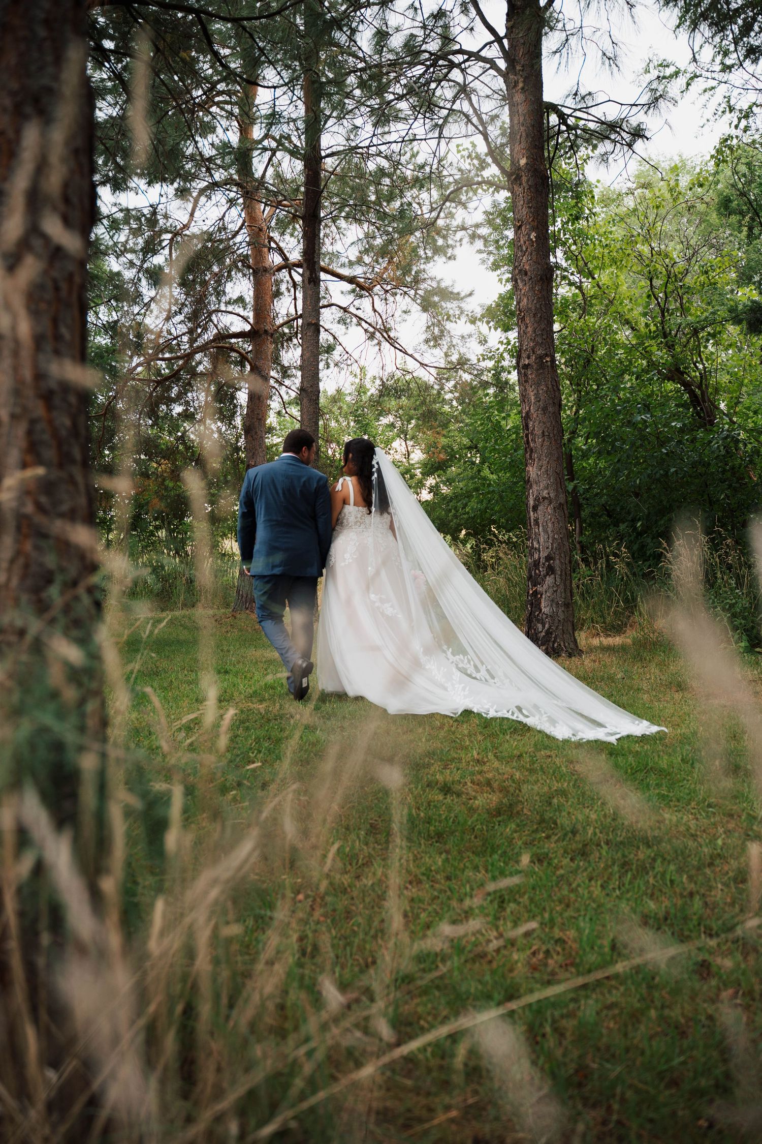 A bride and groom walk hand in hand through a lush green forest, her long white veil trailing behind.