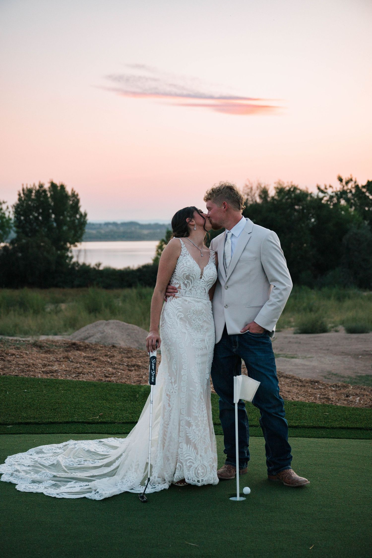 Bride and groom share a romantic kiss on a golf course at sunset, bride in lace gown, groom in white blazer.