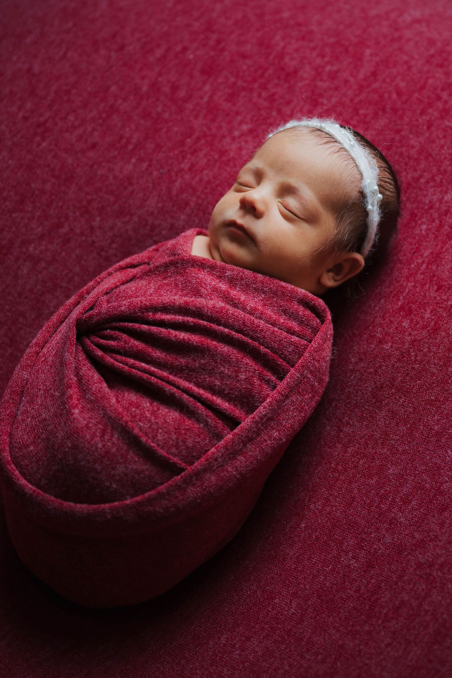 Newborn baby swaddled in a red wrap, sleeping peacefully on a red background wearing a white headband.
