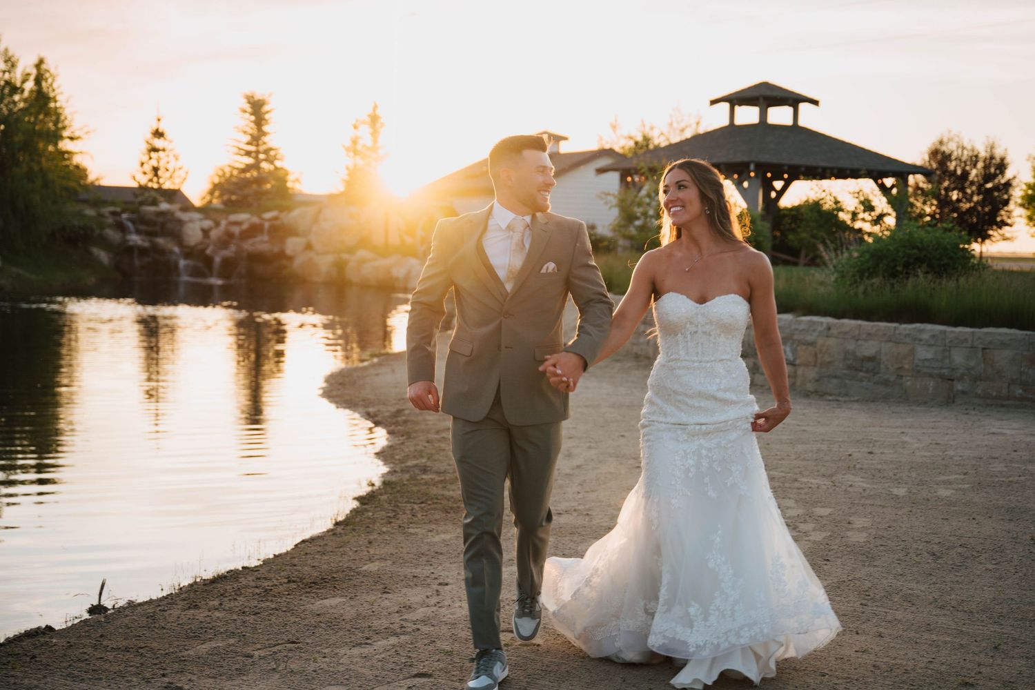 Bride and groom walk hand-in-hand by a pond at golden sunset, with a gazebo in the background at Mint Barrel Barn in Nampa, Idaho