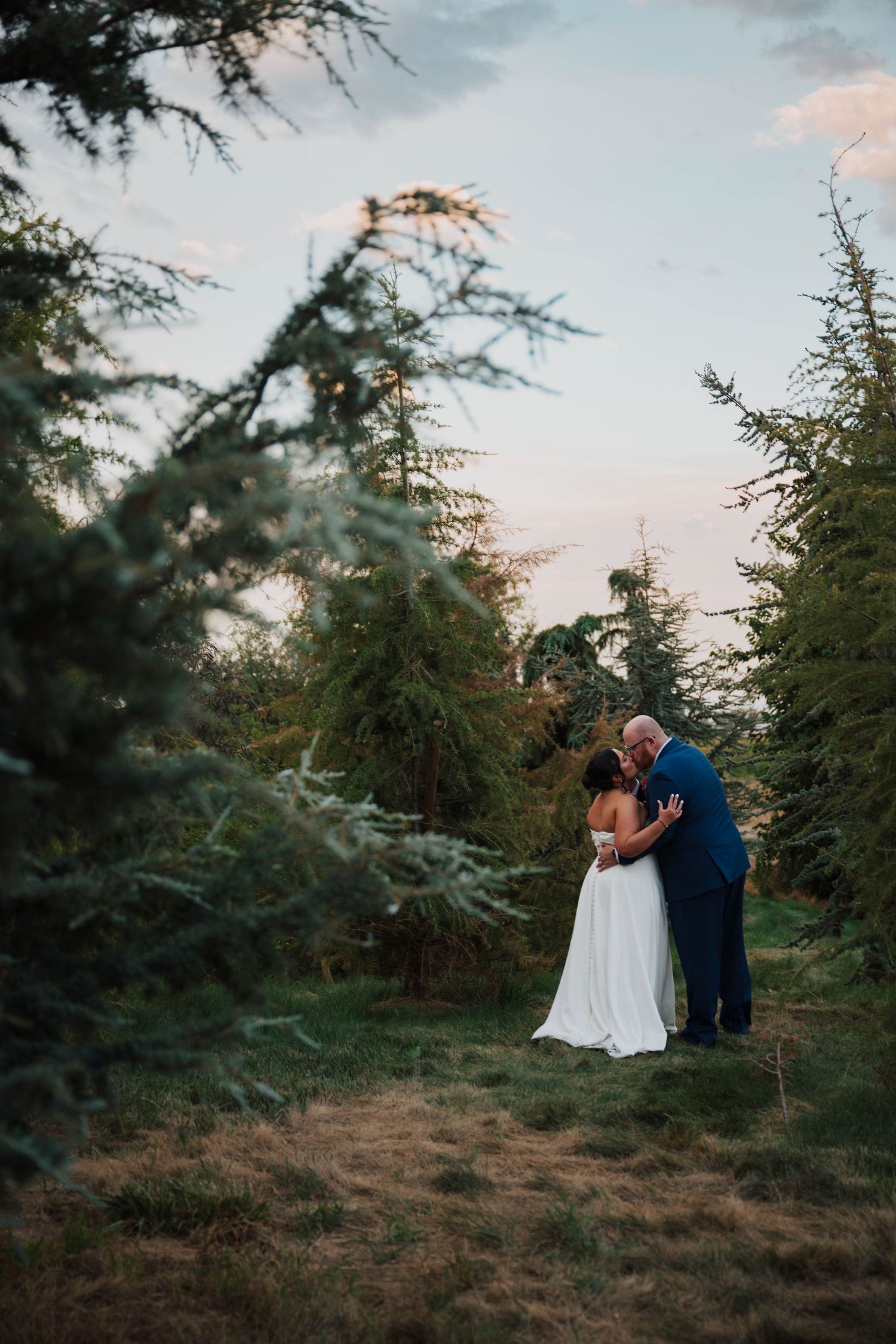 Bride and groom share a romantic kiss surrounded by lush evergreen trees at golden hour in an outdoor wedding setting.