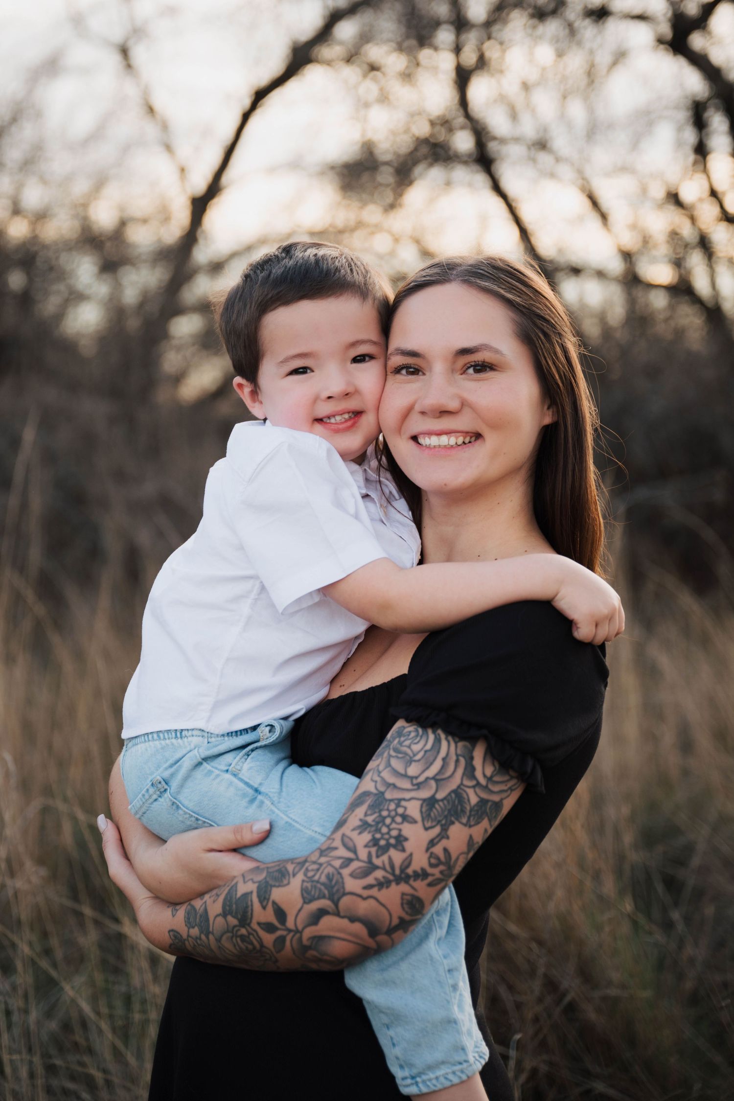 Smiling woman with floral arm tattoo holds a happy young boy outdoors in a golden field at sunset.