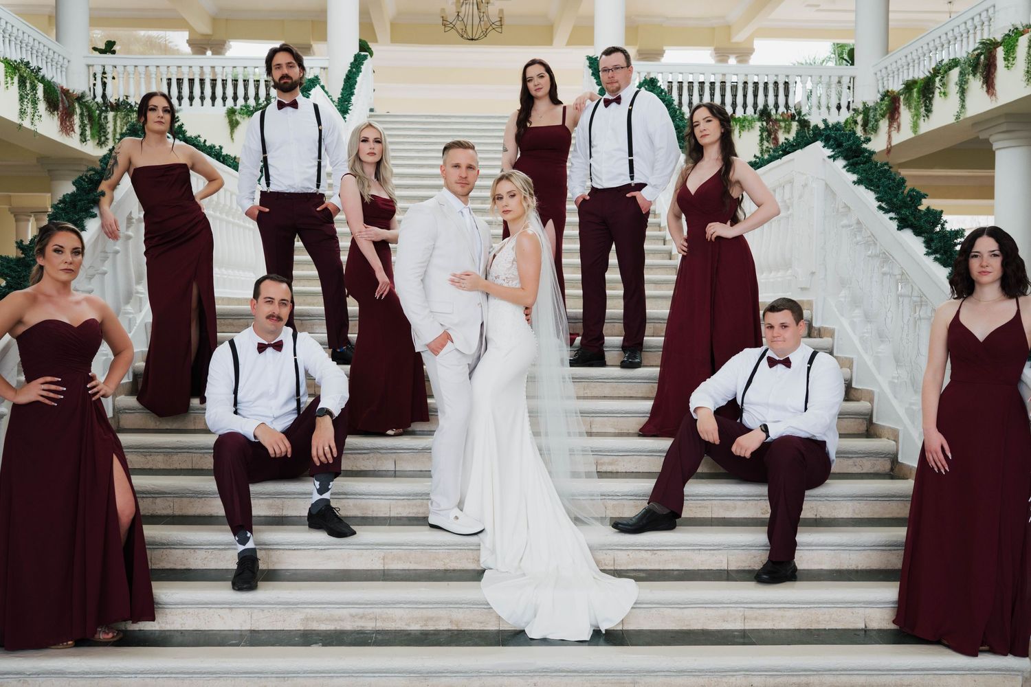 Wedding party poses on grand staircase, bride in white gown with groom, bridesmaids in burgundy dresses, groomsmen in suspenders.