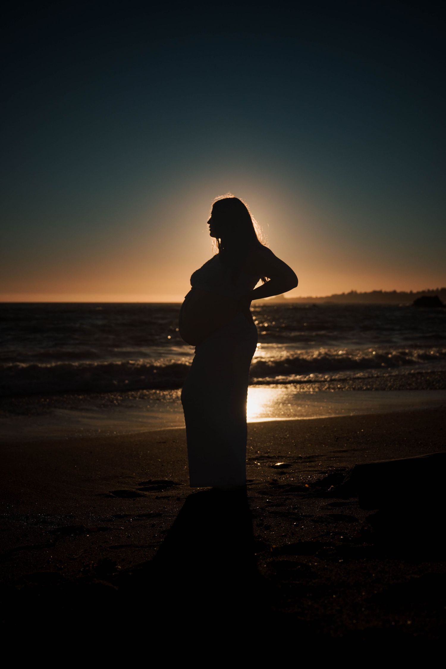 Silhouette of a pregnant woman standing on a beach at sunset with ocean waves in the background.