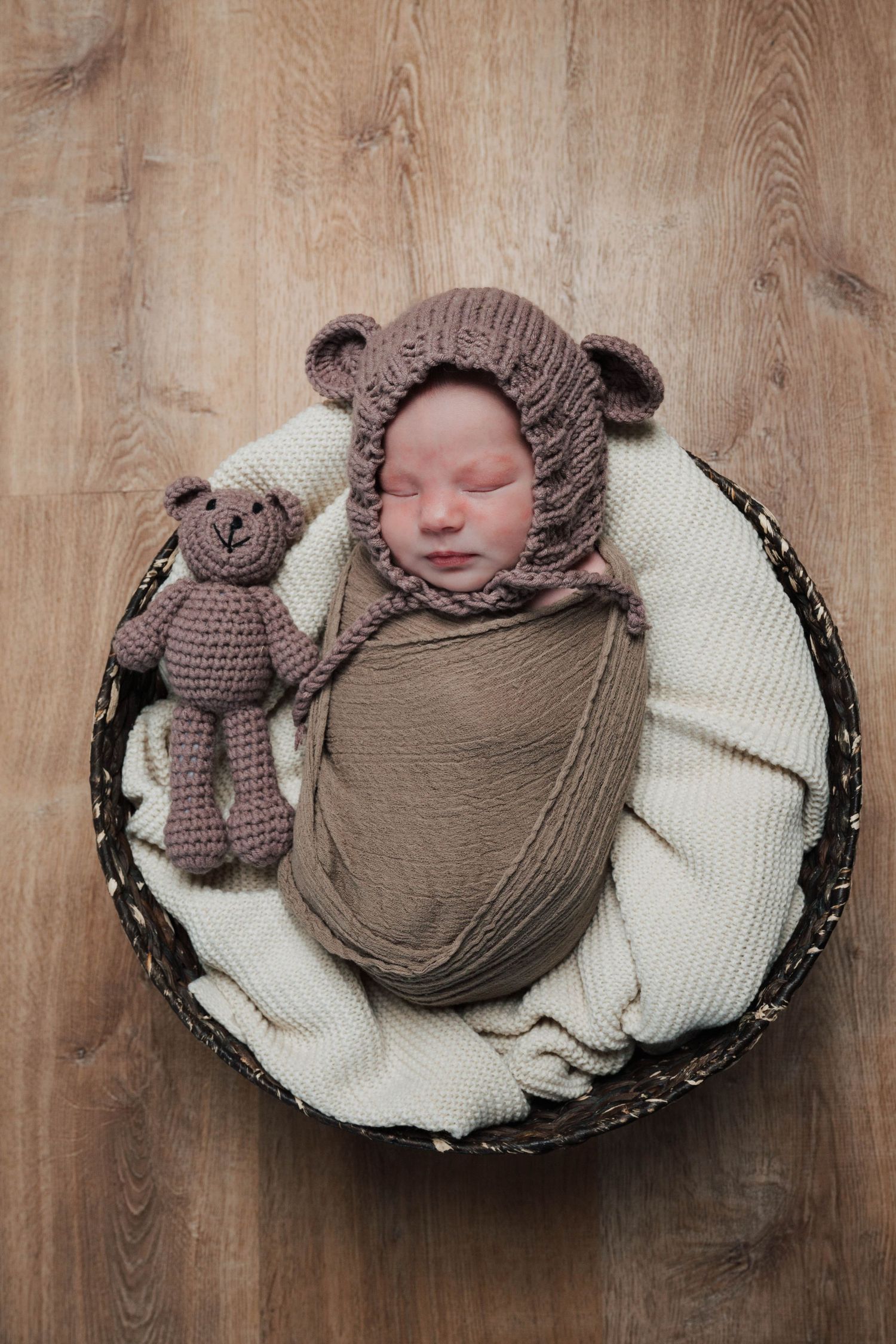 Newborn baby in brown wrap with teddy bear prop in rustic basket on wooden floor.