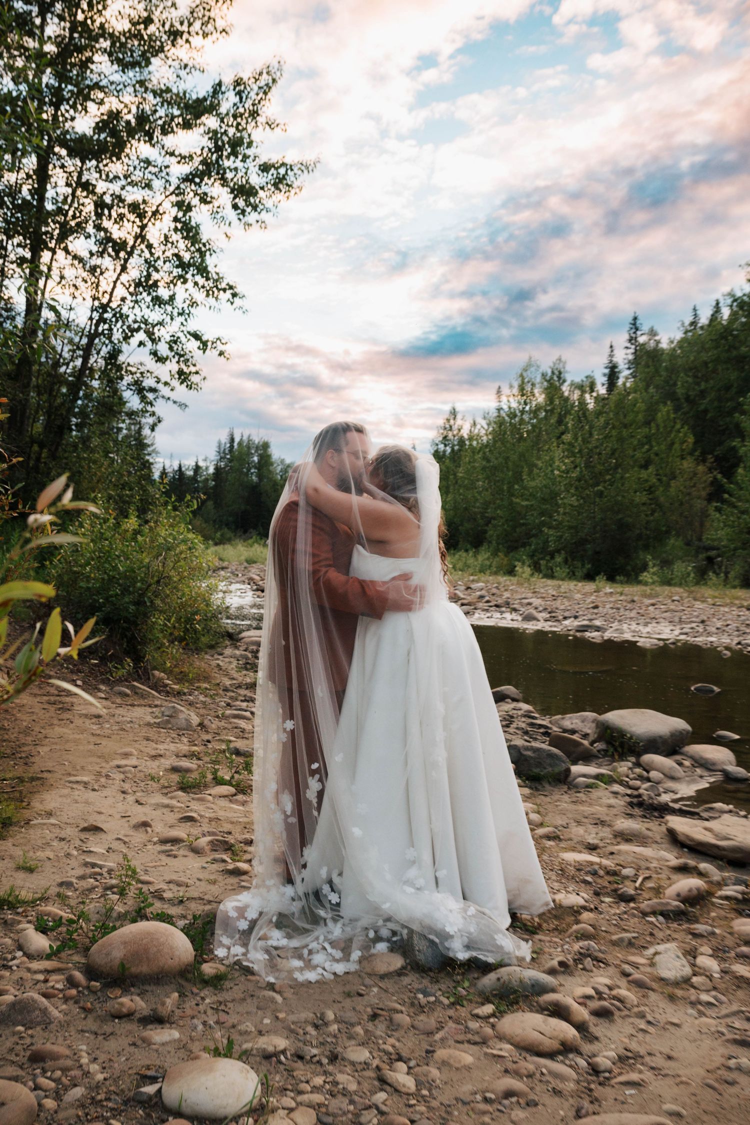 Two brides embrace near a rocky riverbank surrounded by lush forest under a pastel sunset sky.