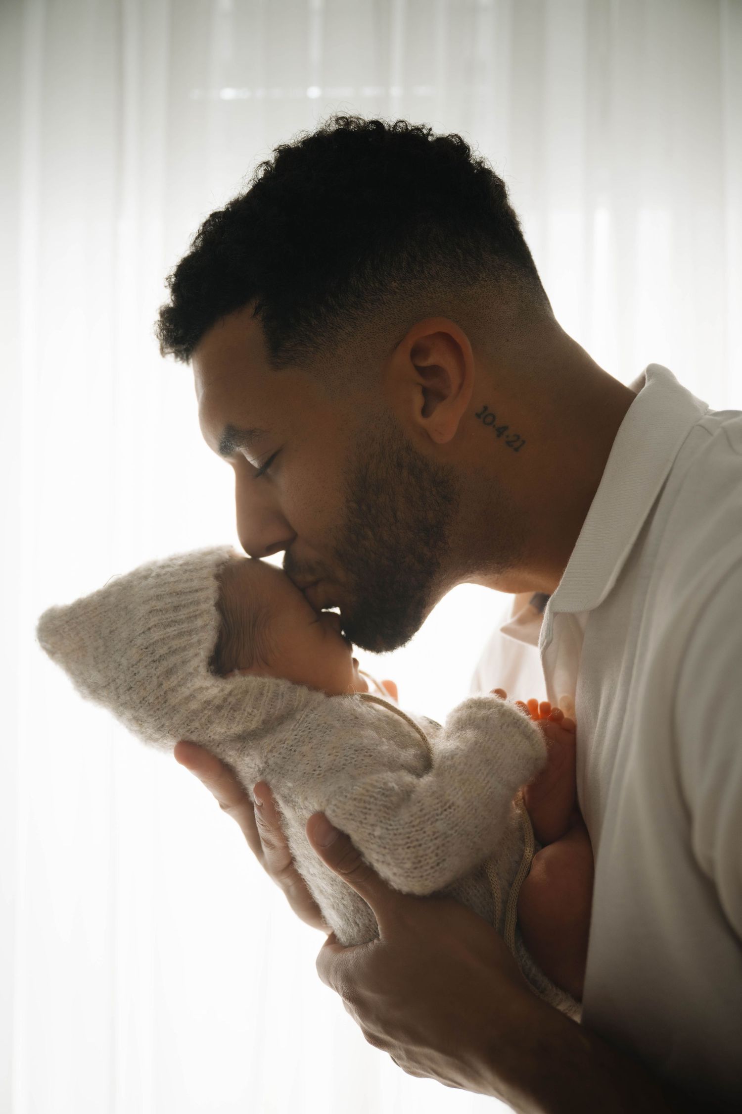 A father gently kisses his newborn baby wrapped in a white knit blanket near a bright window.