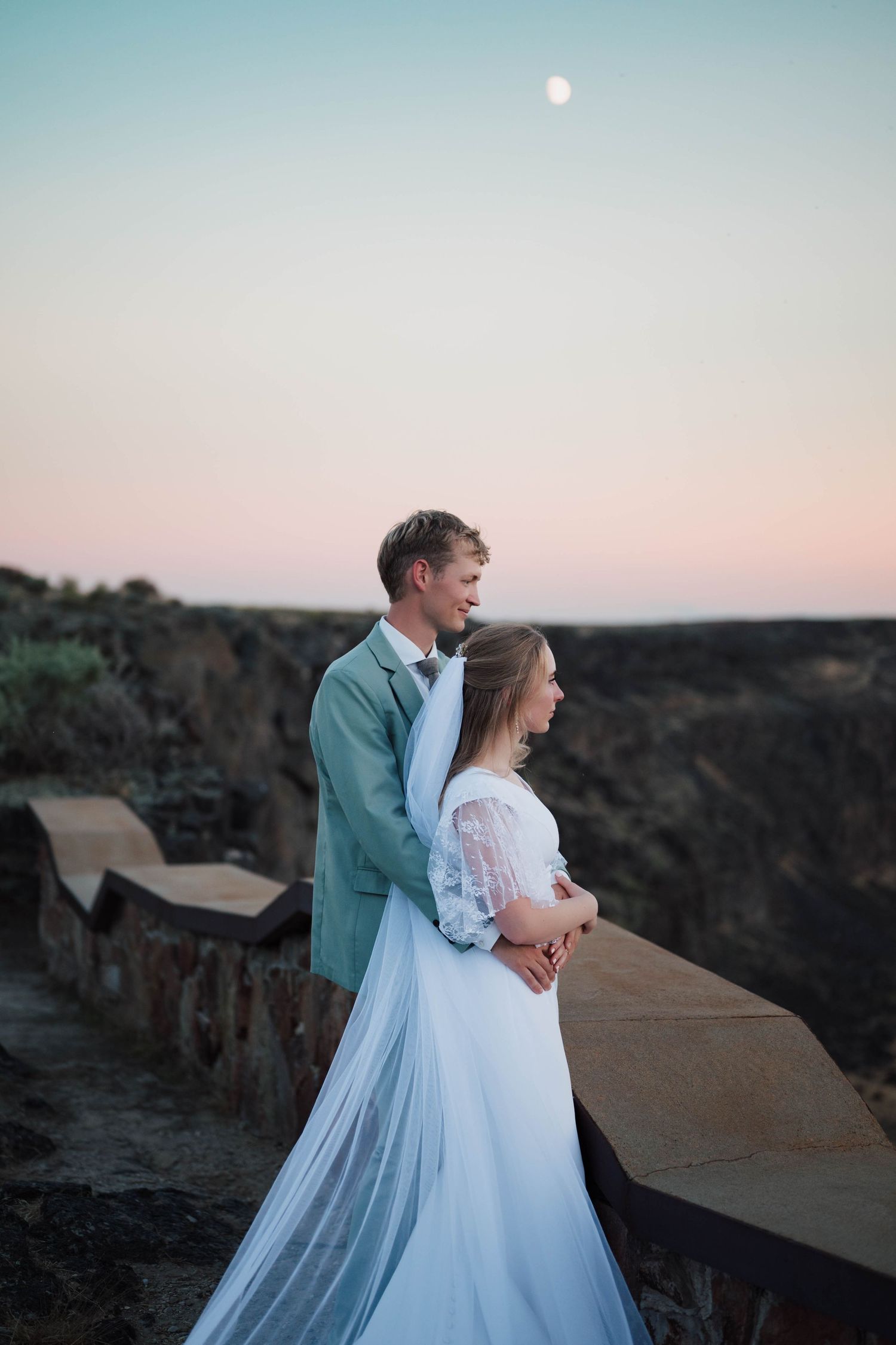 Bride and groom embrace at dusk on a rocky overlook with a full moon rising in the pastel sky.