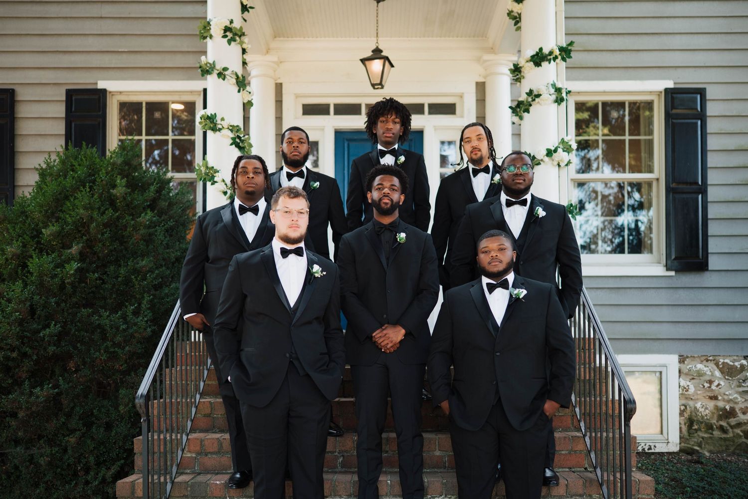 Groomsmen in black tuxedos and bow ties pose on steps of elegant home entrance for wedding photo.