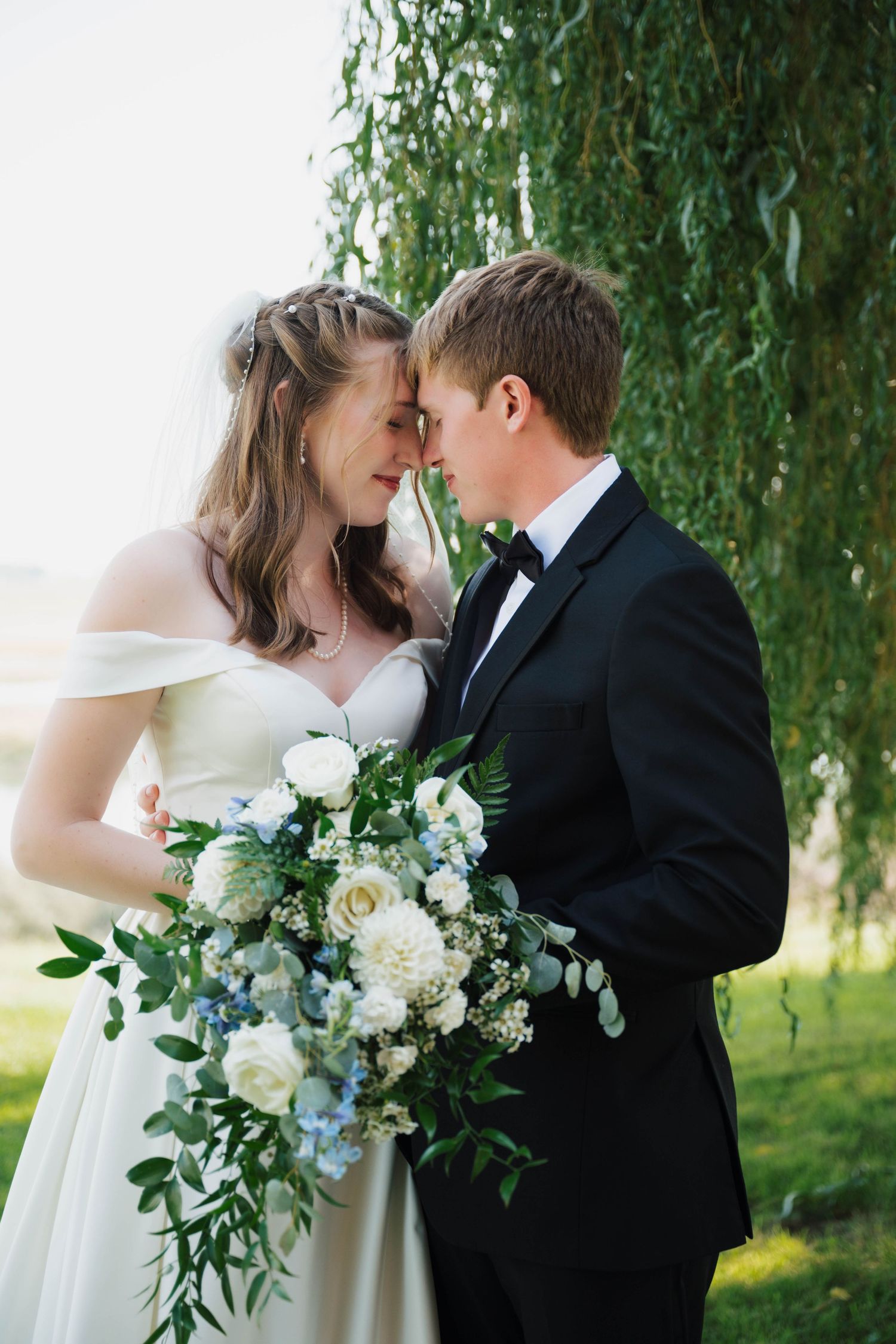 A romantic moment captured under a weeping willow tree with a white floral bouquet.