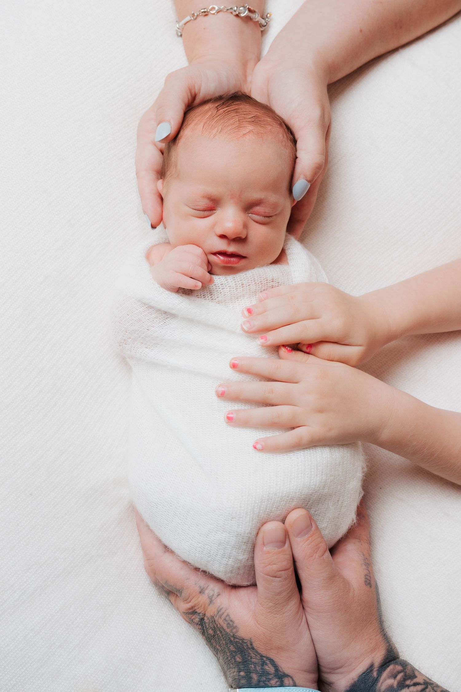 Newborn baby swaddled in white, cradled by parents hands, one with tattoos and one with a charm bracelet.