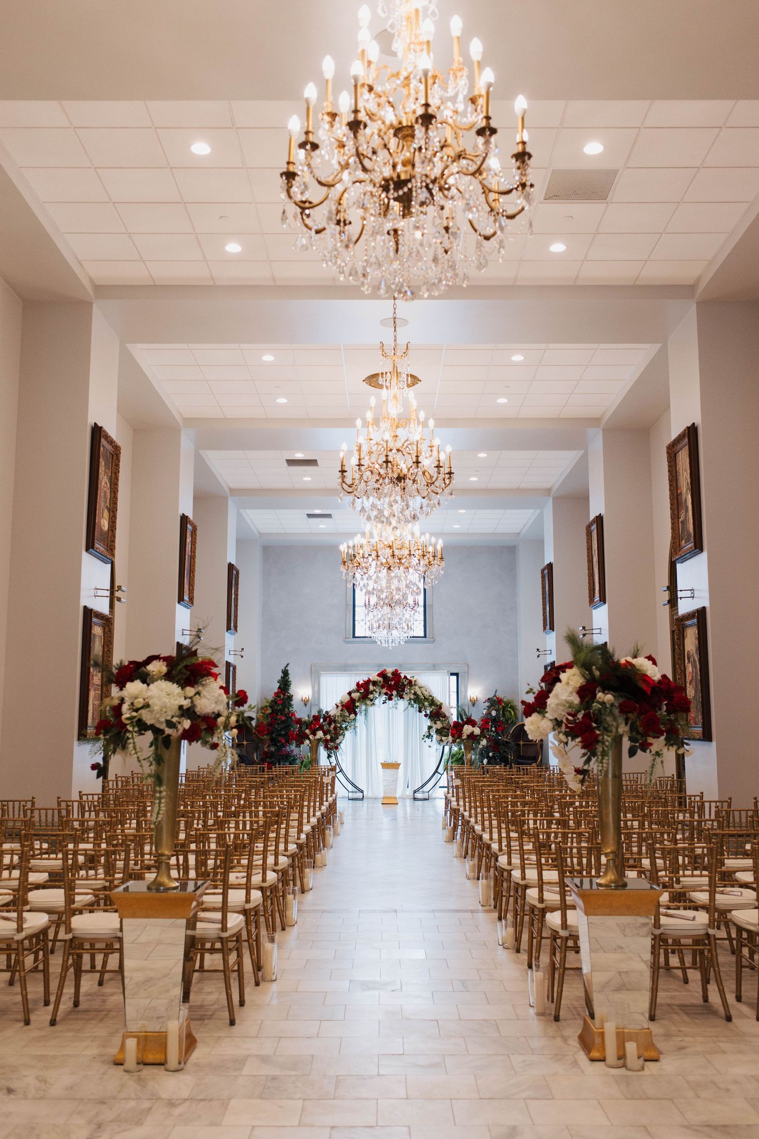 Elegant wedding ceremony venue with crystal chandeliers, gold chairs lining the aisle, and red and white floral arrangements.
