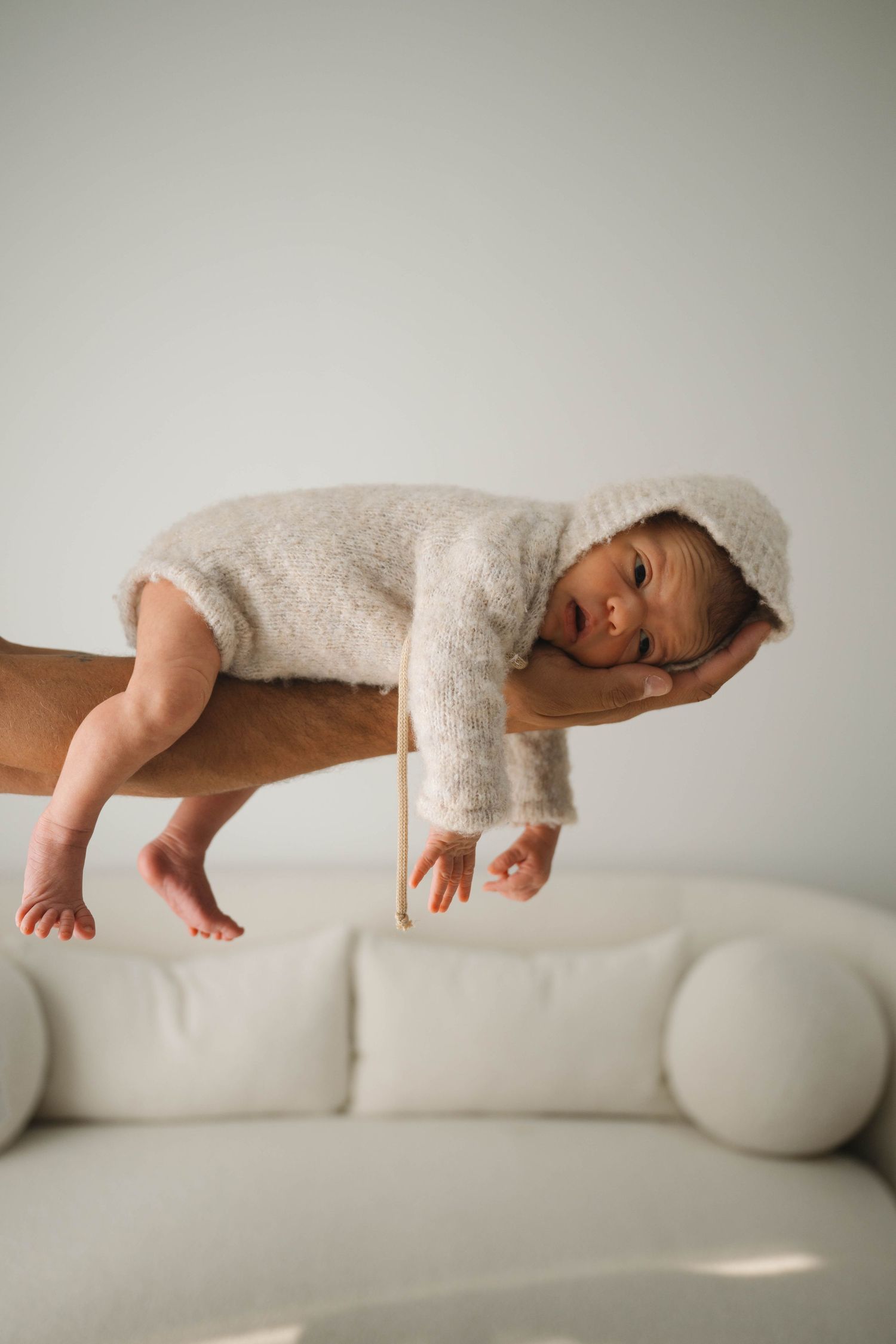 Newborn baby in fuzzy hooded onesie resting on adult hand above white sofa.