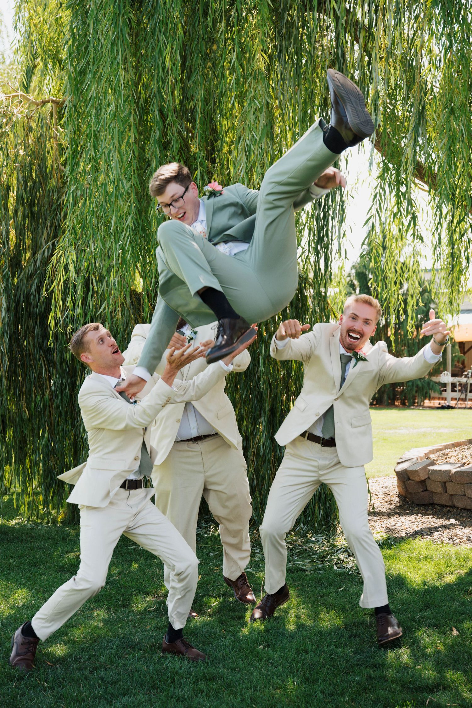Groomsmen in light colored suits performing playful jumping poses under a weeping willow tree.