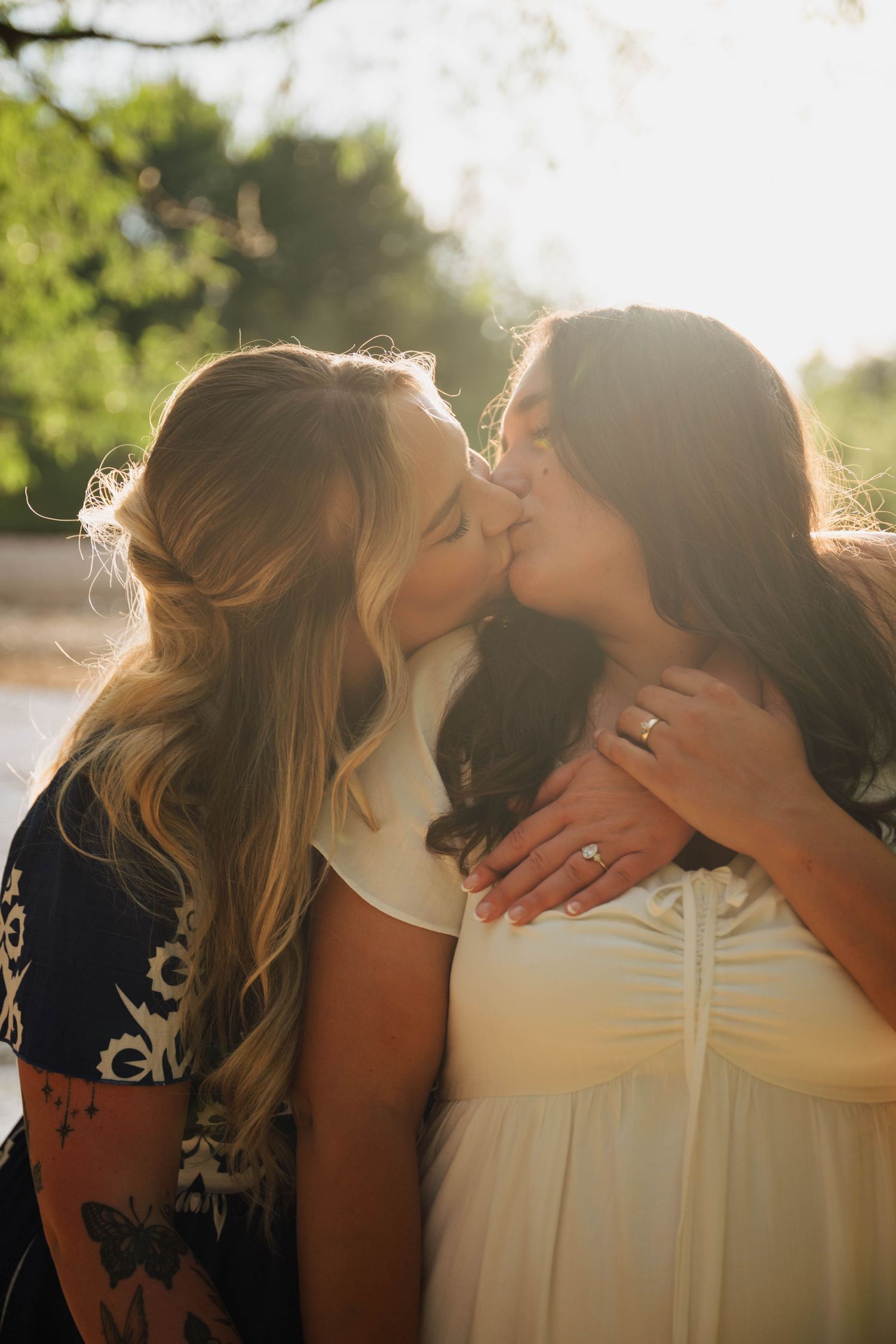 Two women sharing a romantic kiss outdoors, one wearing a white dress with an engagement ring, backlit by golden sunlight.