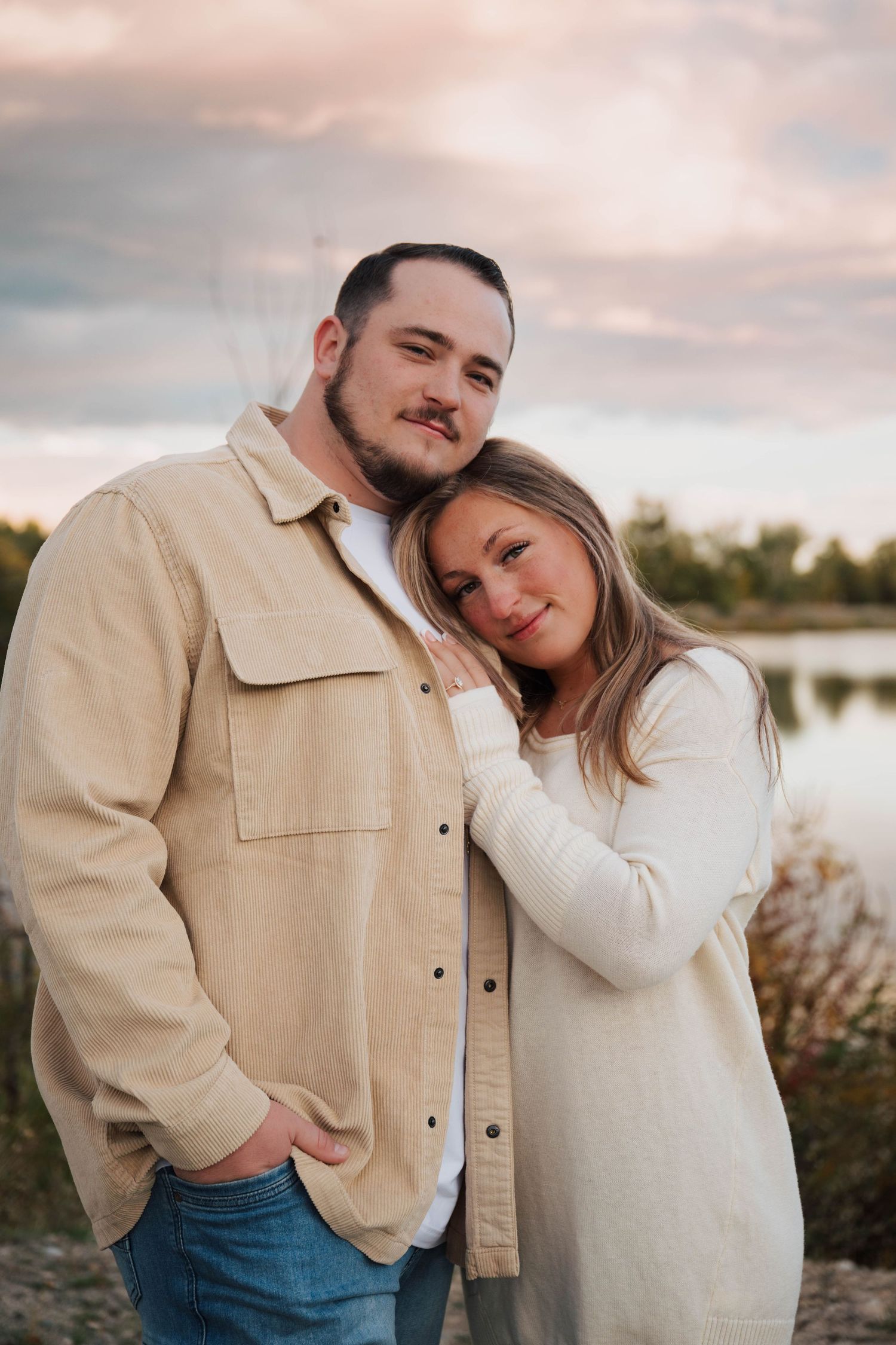 A couple in casual attire shares an affectionate moment by a lakeside during sunset.