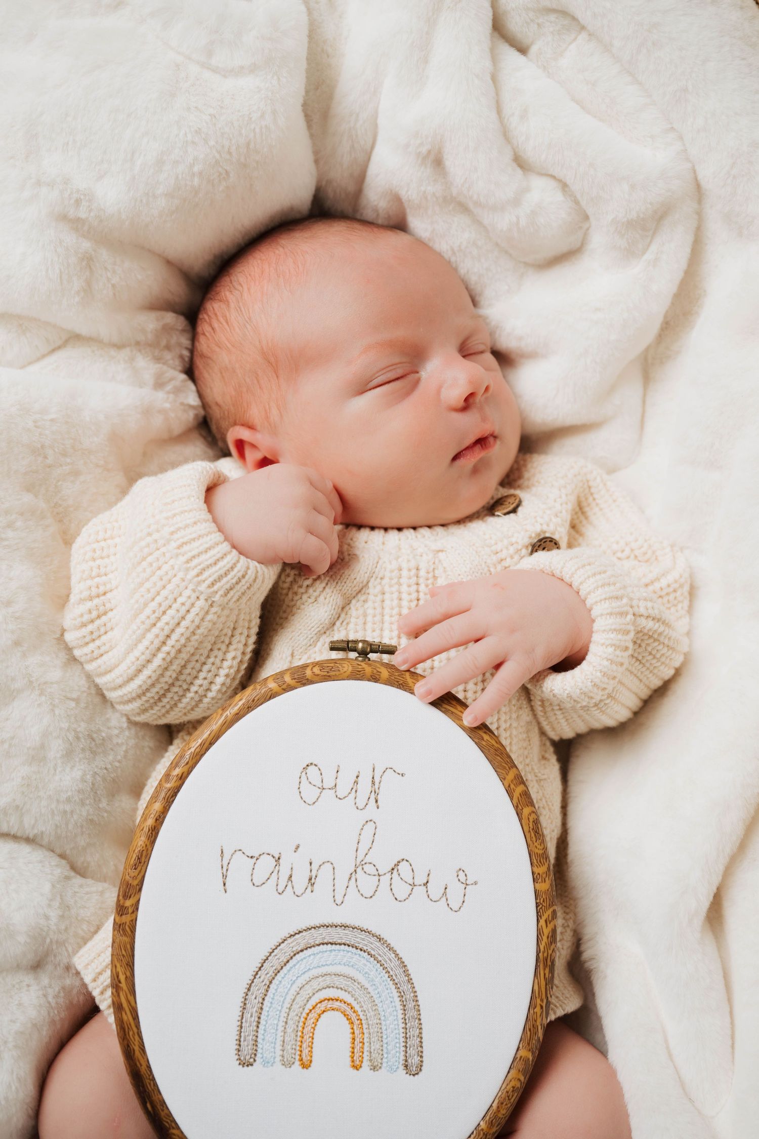 Sleeping newborn in cream sweater with embroidery hoop showing rainbow design.