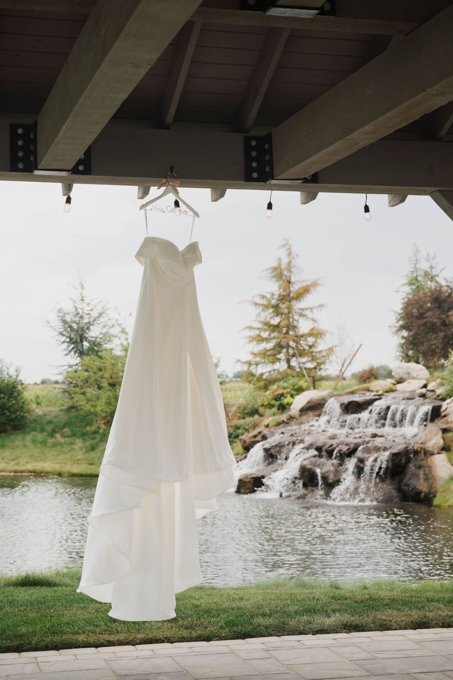 White strapless wedding dress hanging on wooden beam with waterfall and pond in background.