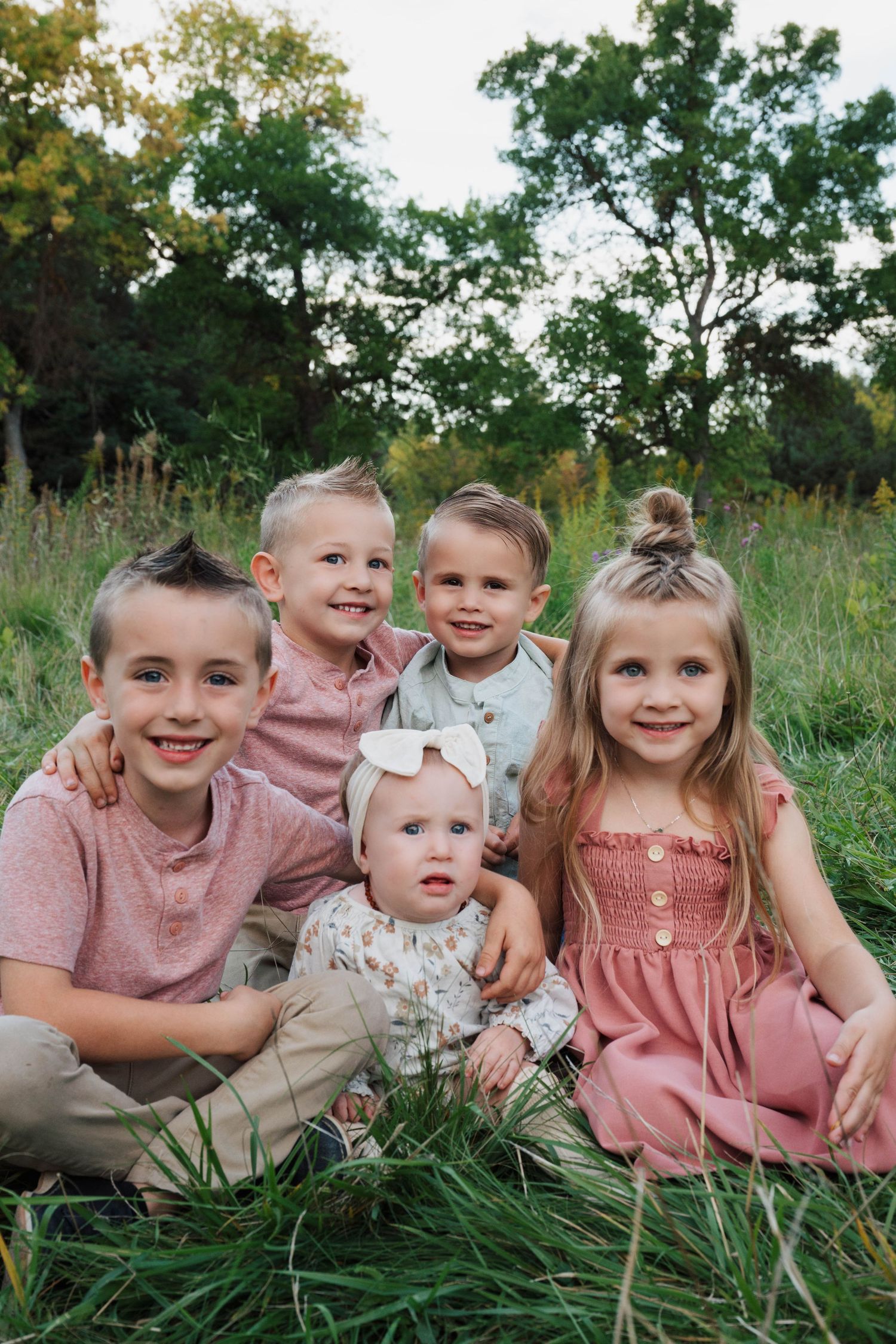 Young children in pink and neutral colored outfits sit together in tall grass during a summer photoshoot.