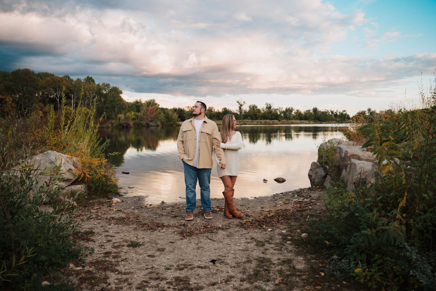 A couple shares an intimate moment by a tranquil lake during sunset with trees in the background.