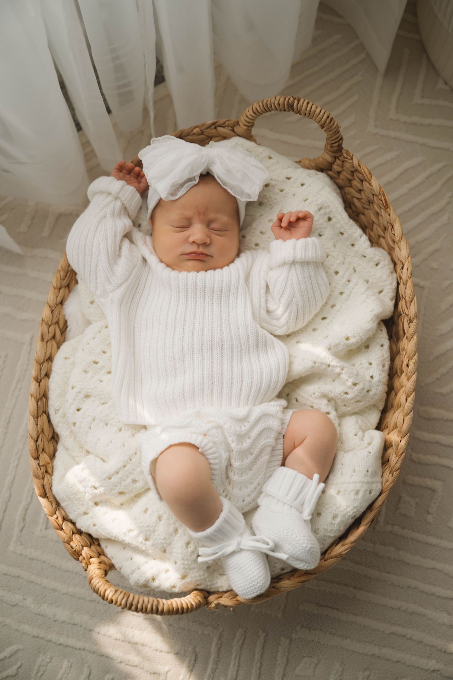 Sleeping newborn baby in white knit outfit and bow headband resting in a woven basket with cream blanket.