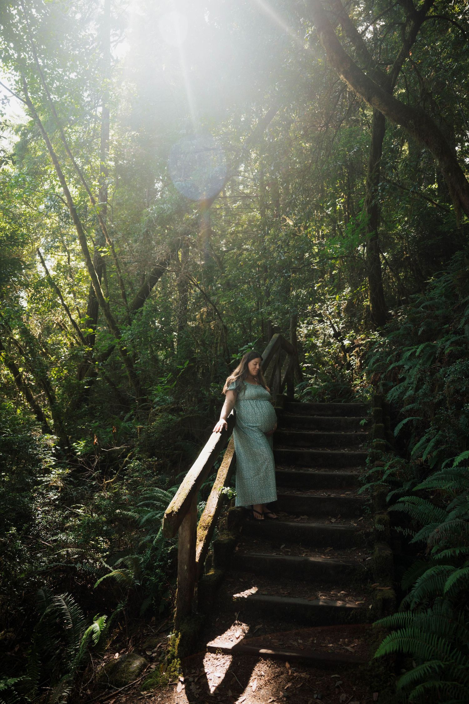 Pregnant woman in mint dress standing on forest stairs surrounded by lush ferns with sunlight filtering through trees.