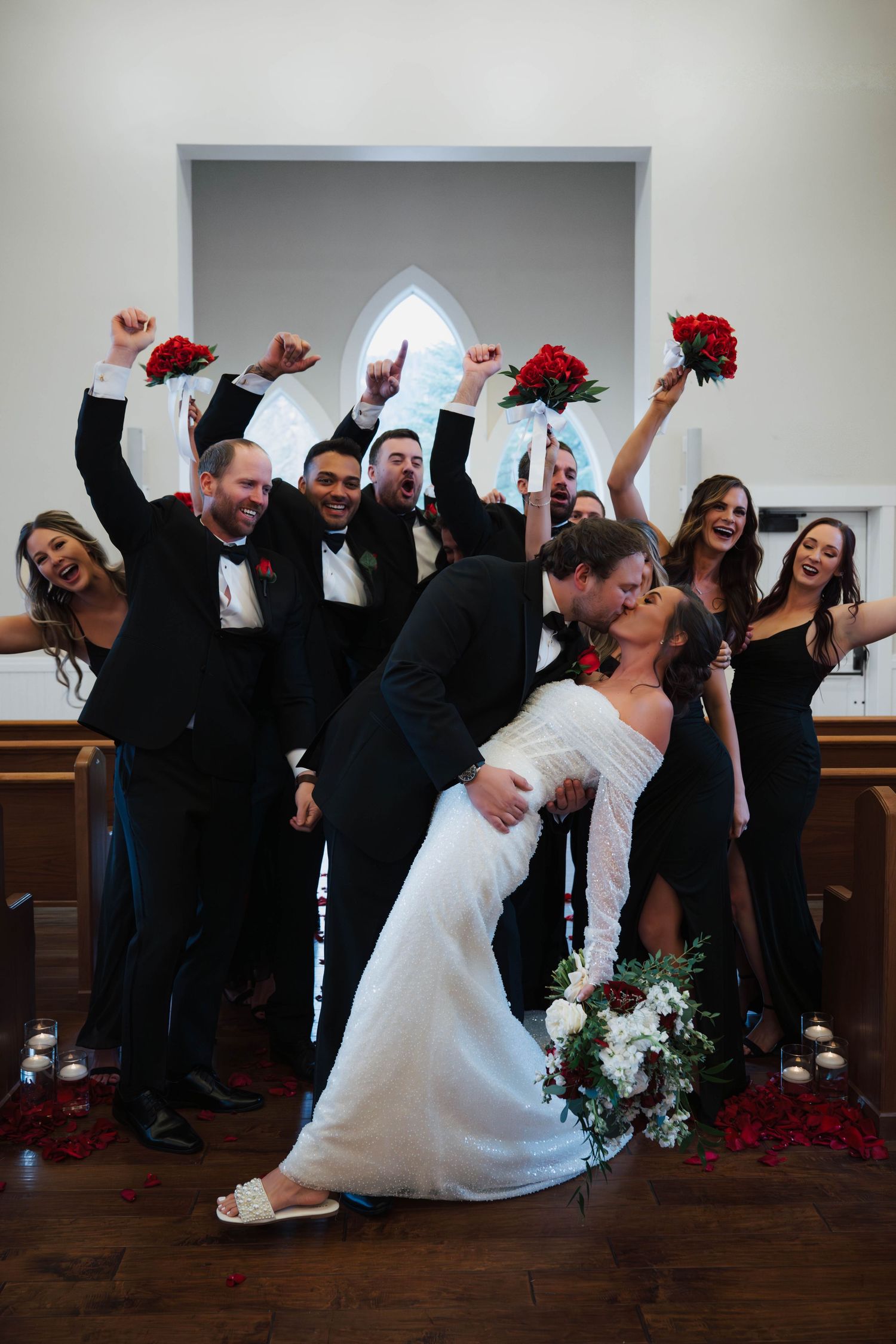 Bride and groom kiss as excited wedding party celebrates behind them in a church with red roses.