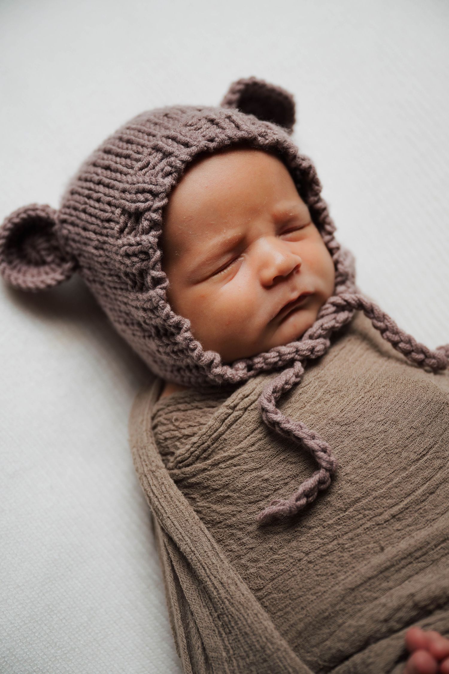 Newborn baby sleeping peacefully swaddled in brown wrap wearing a cute knitted bear ear bonnet hat.