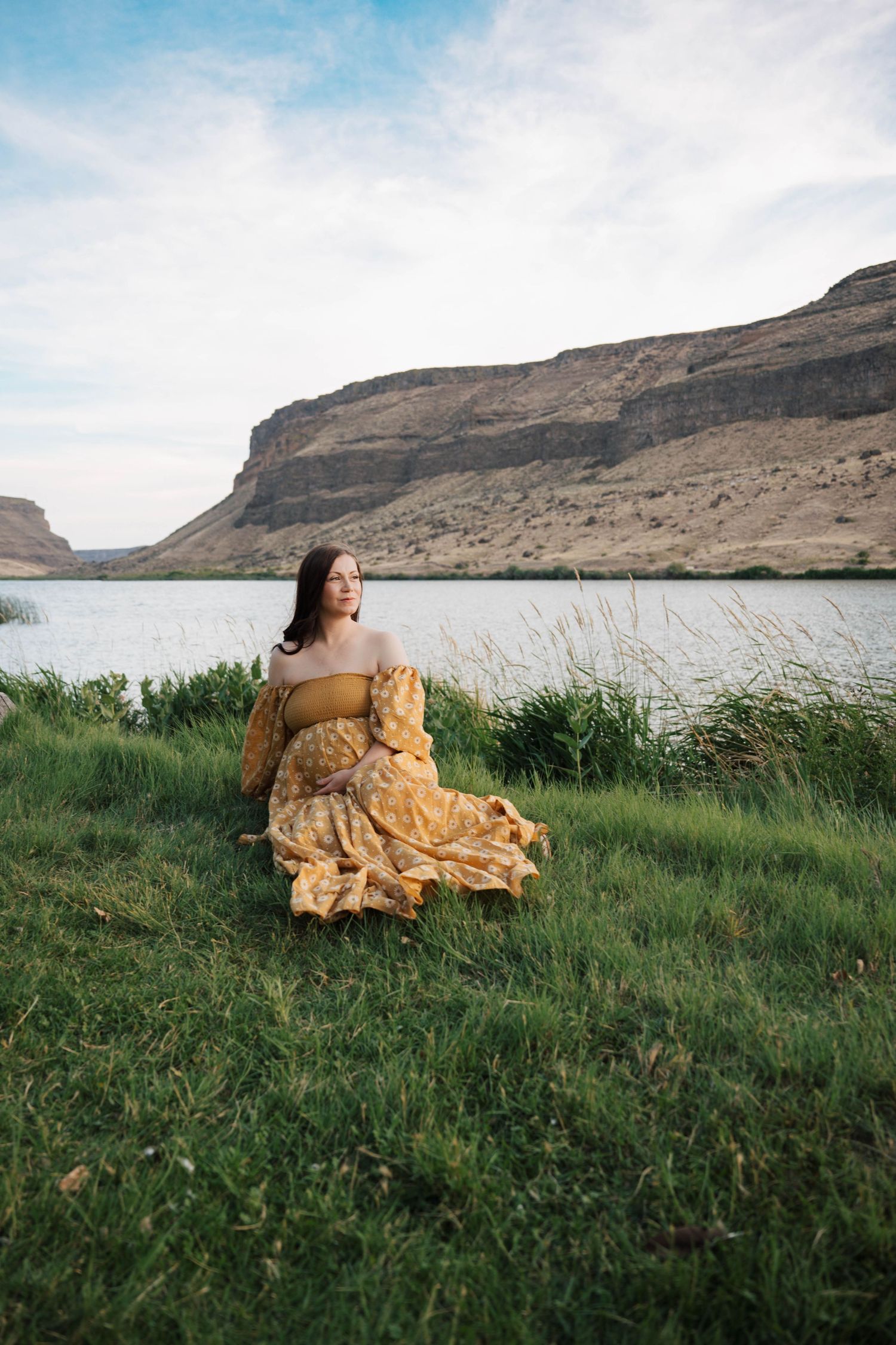 Person in flowing yellow dress poses dramatically on grassy hillside with cliffs in background.