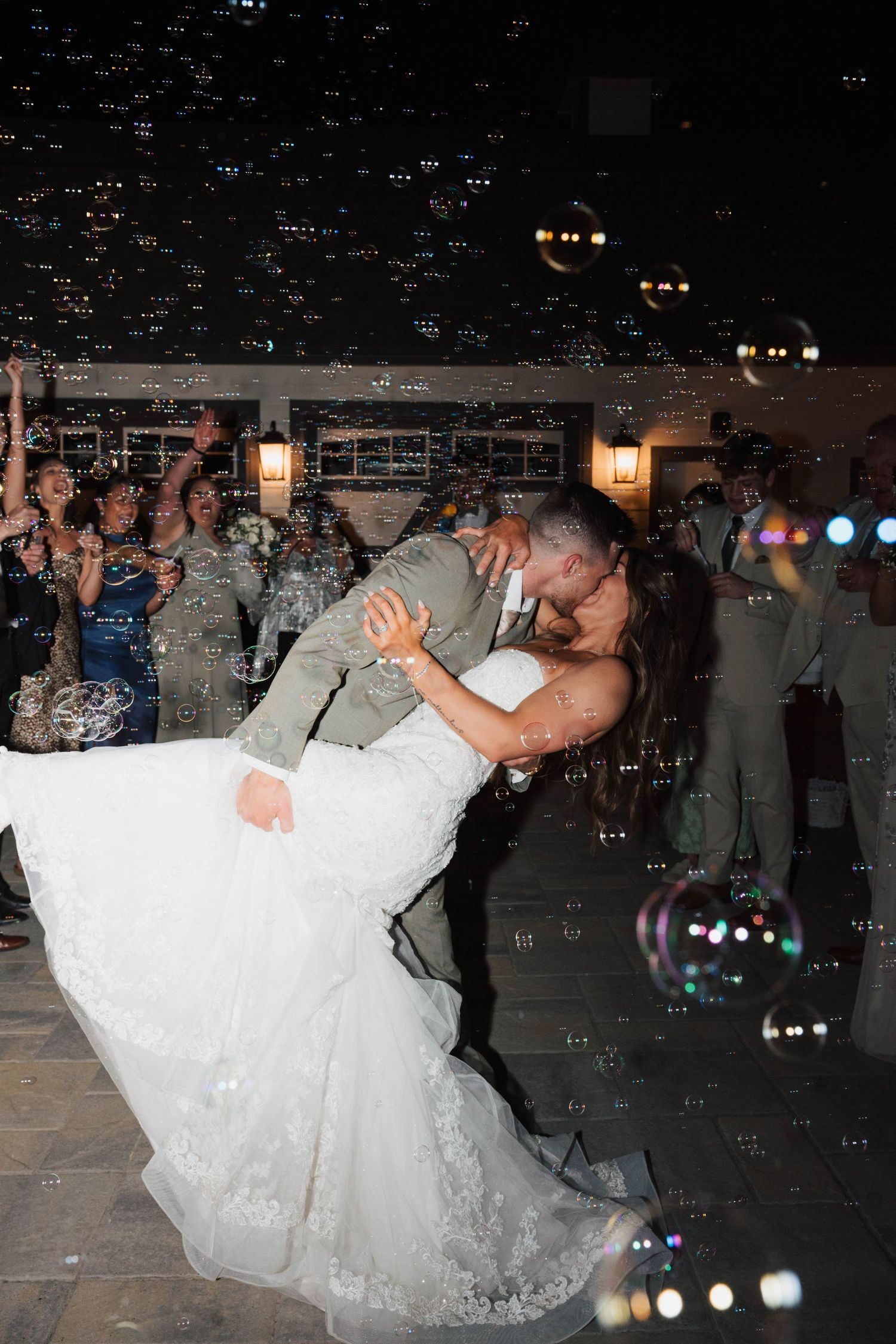 Groom dips bride for a romantic kiss on the dance floor surrounded by bubbles and cheering wedding guests at night.
