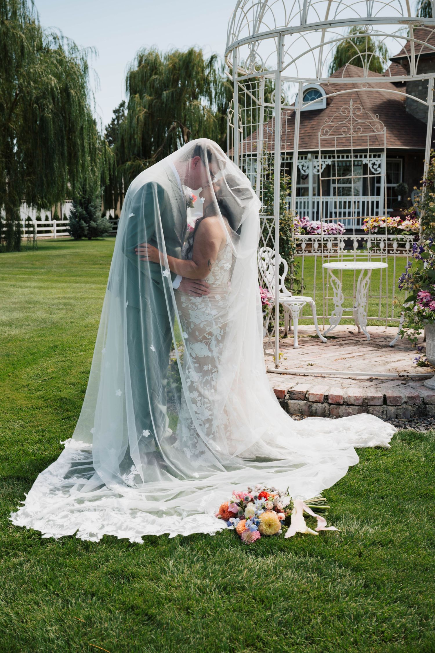 Bride in lace gown posing gracefully on grass near white garden gazebo with flowing veil.