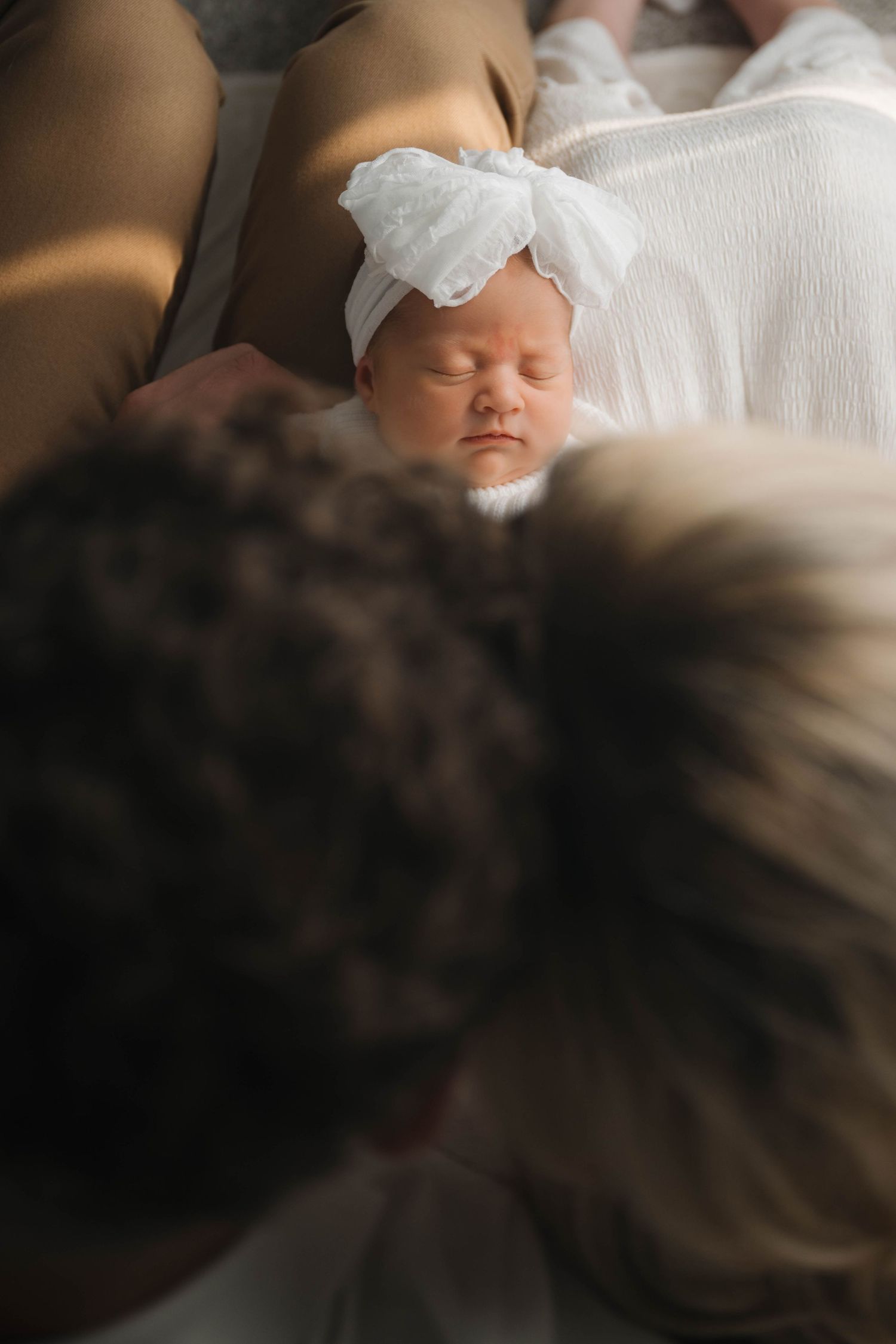 Newborn baby girl sleeping peacefully in white swaddle with bow headband, cradled by parents.