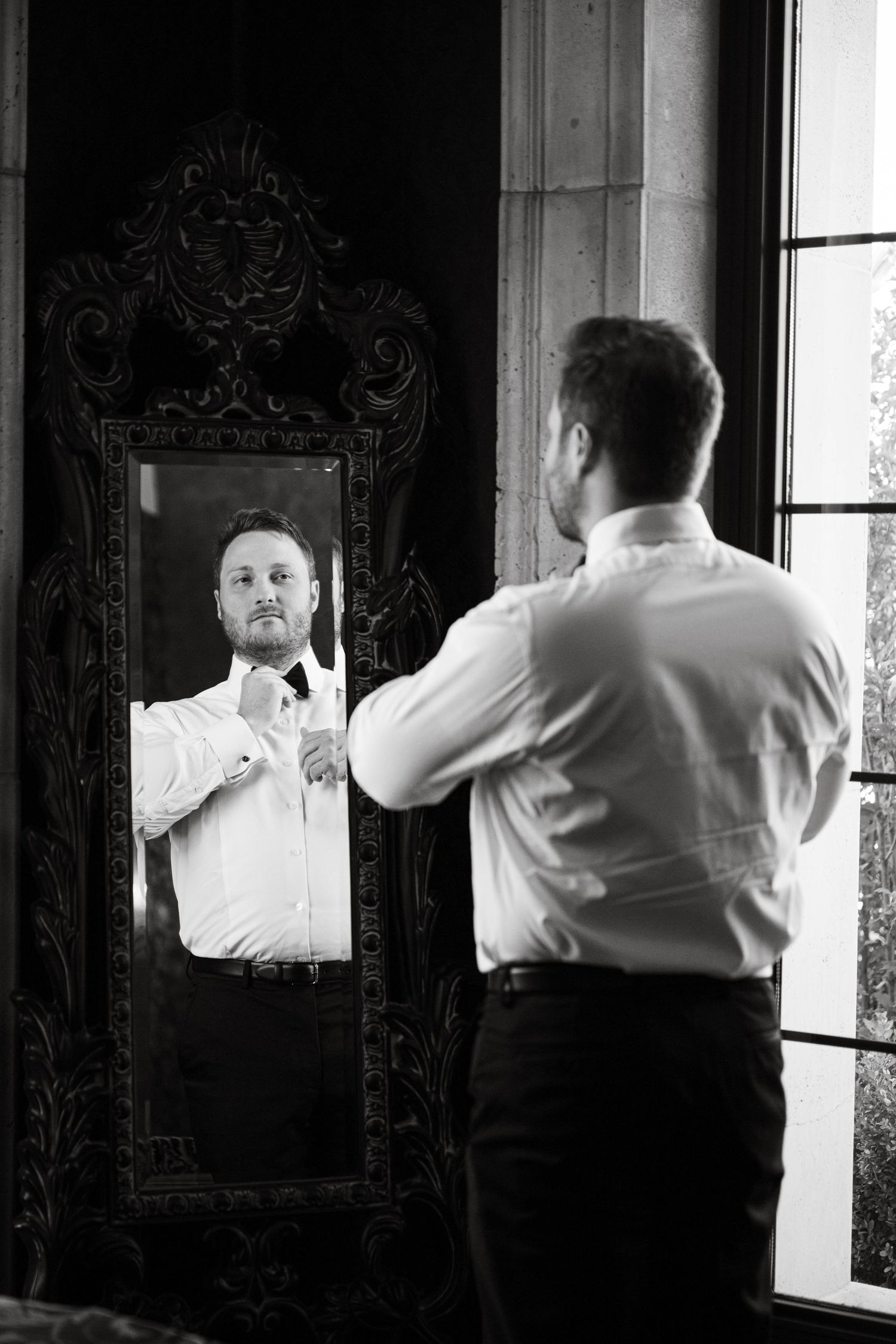 Black and white photo of a groom adjusting his bow tie while looking in an ornate mirror before his wedding.