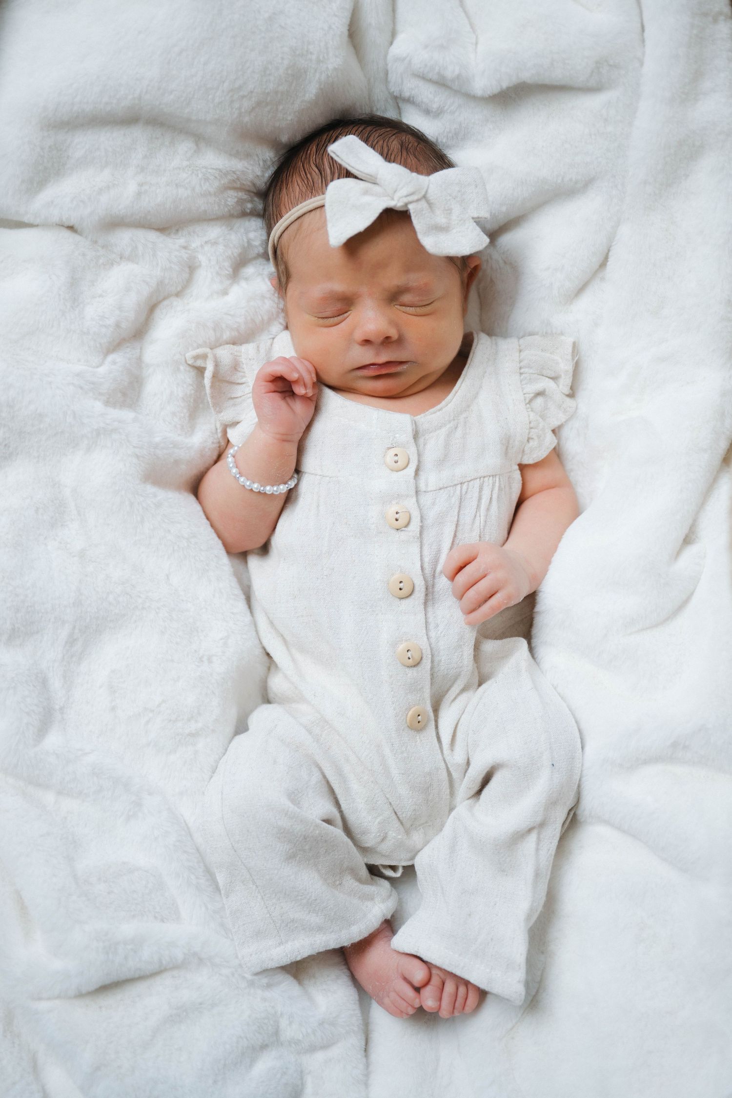 Sleeping newborn baby girl in white linen romper and bow headband lying on soft white blanket.