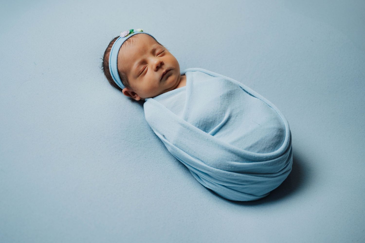 Newborn baby swaddled in light blue wrap with floral headband, sleeping peacefully on blue background.