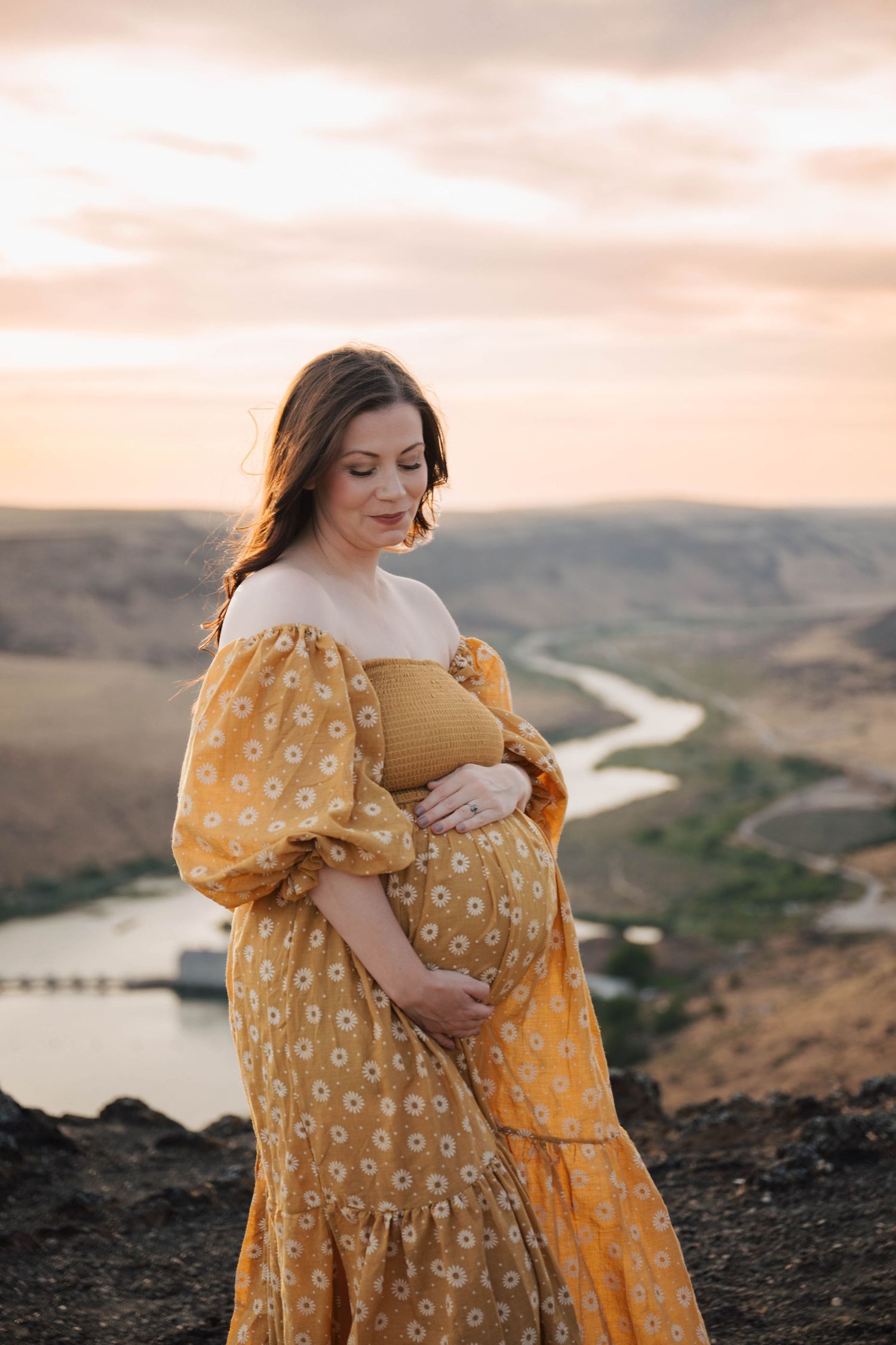 Pregnant woman in yellow floral dress stands on cliff overlooking winding river during sunset.