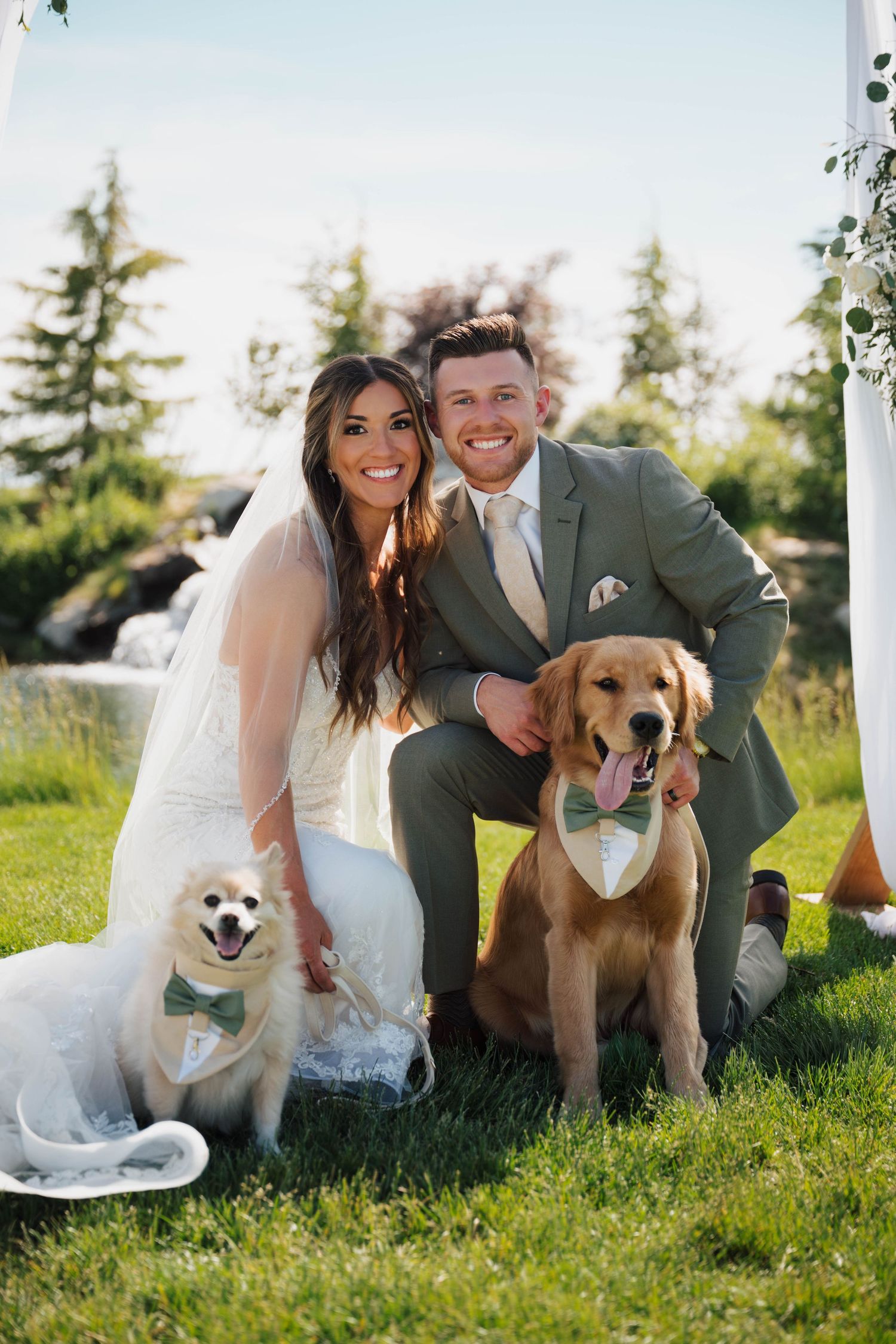 Smiling bride and groom posing with two dogs wearing bow ties on green grass at outdoor wedding ceremony.