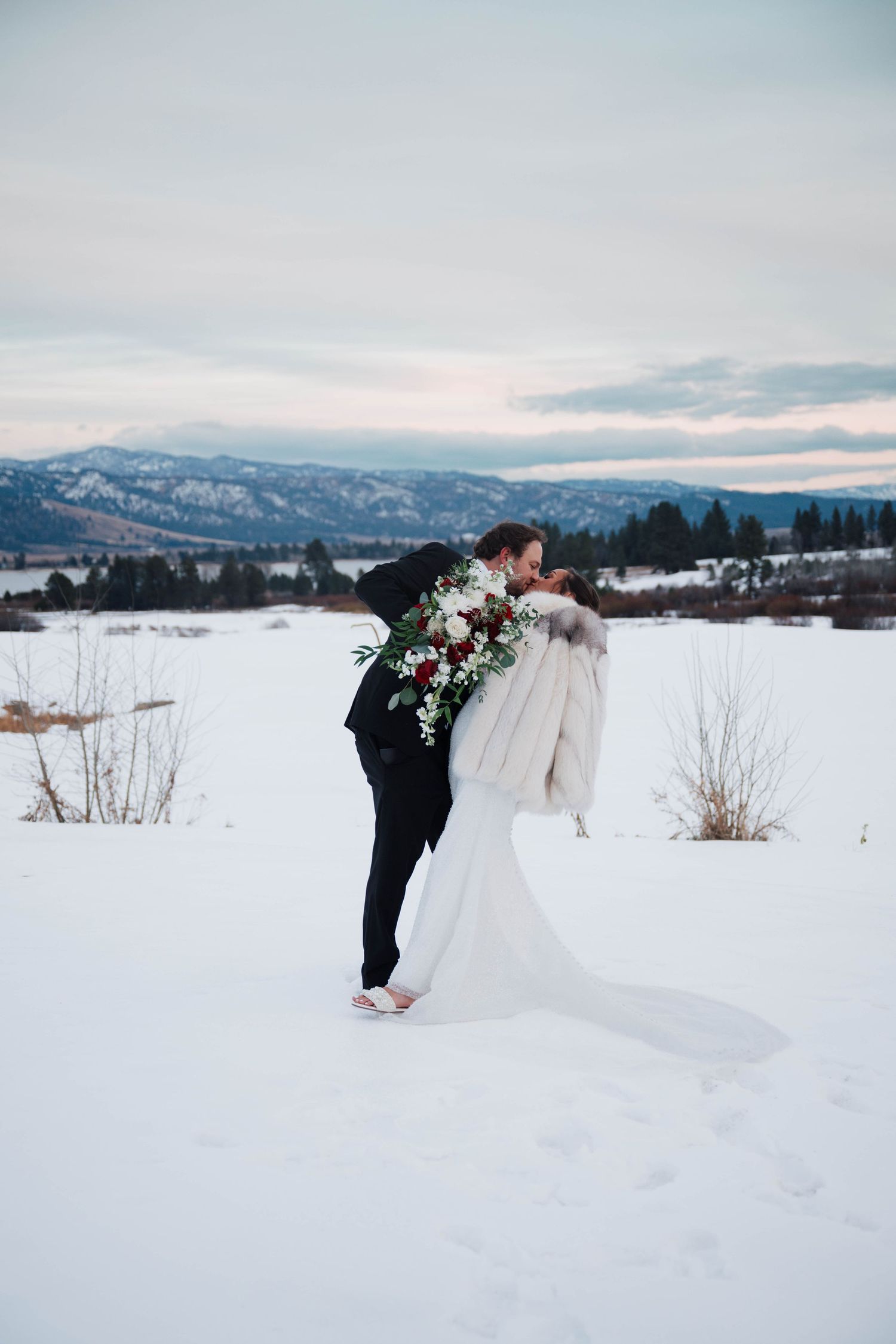 Bride and groom embrace in a snowy winter landscape with mountain views and a red bouquet.