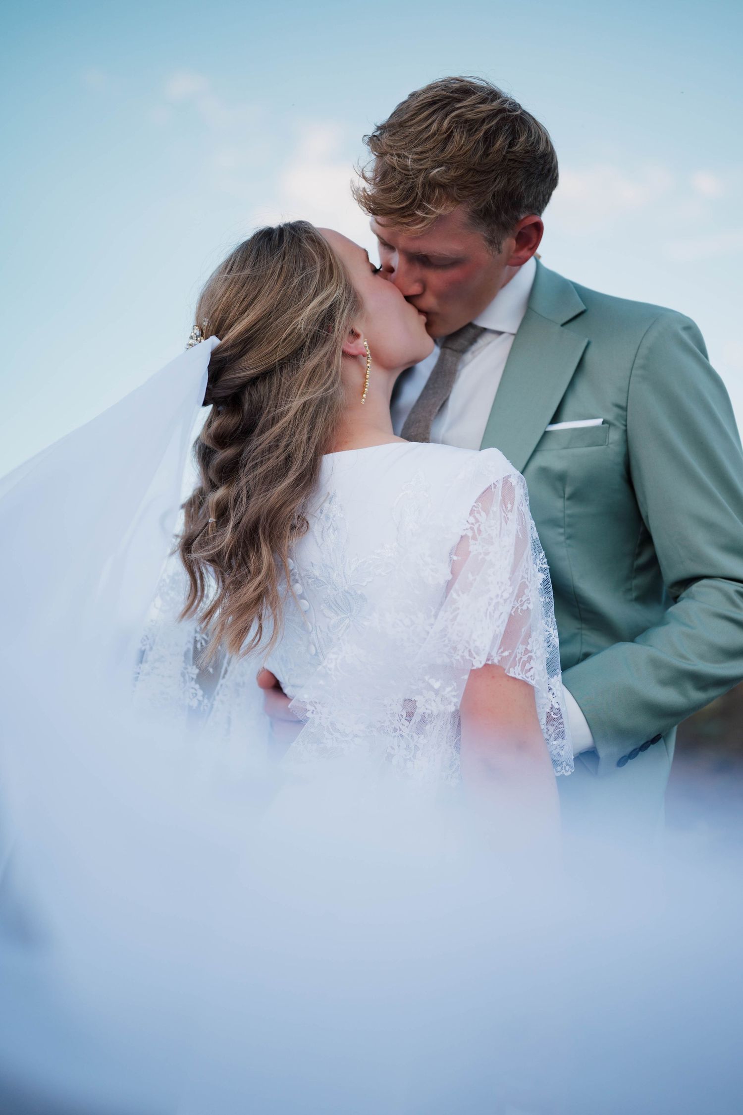 Bride and groom sharing a romantic kiss outdoors, bride in lace dress with veil, groom in sage green suit.