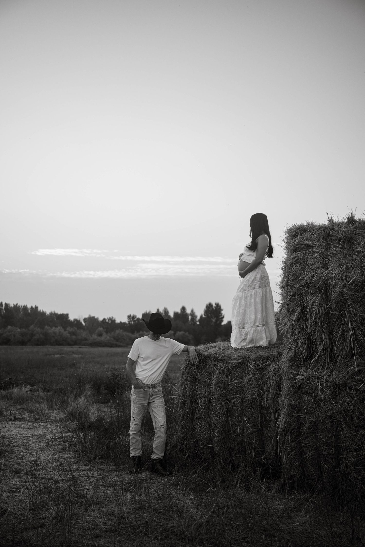 Black and white photo of a boy and pregnant woman by a hay bale in a rural field at dusk.