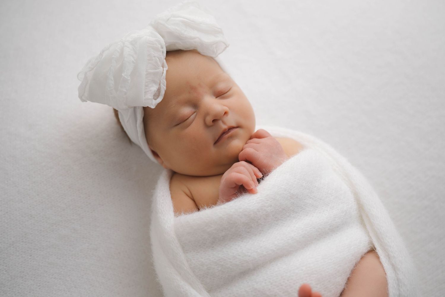 Peaceful newborn baby girl swaddled in white wrap with bow headband, sleeping on white background.