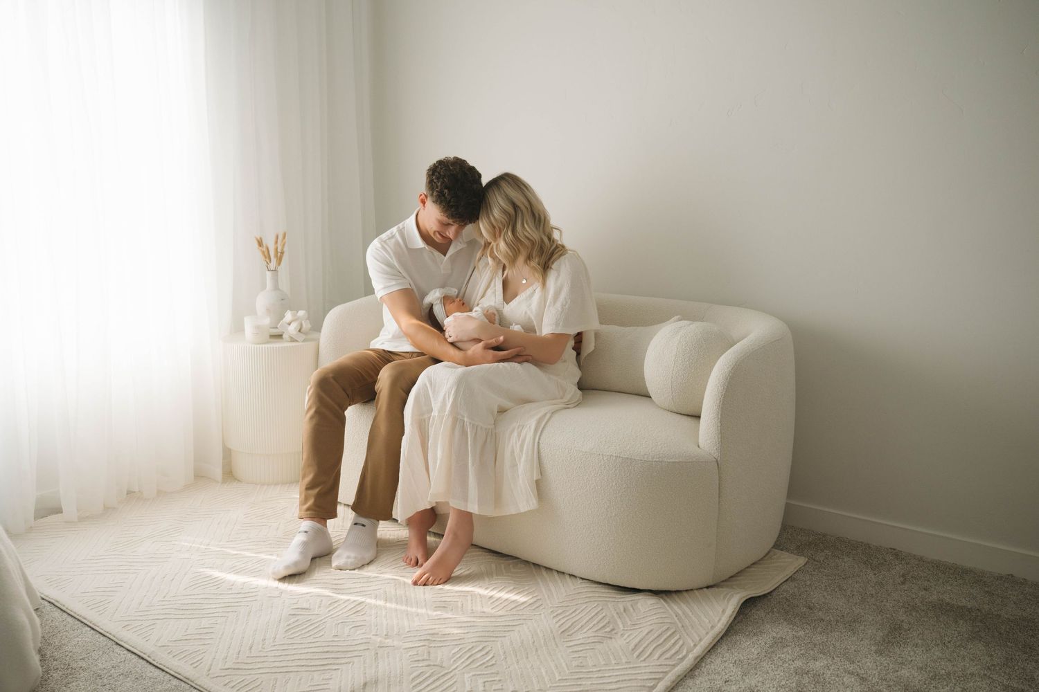 A couple sits on a white sofa, lovingly gazing at their newborn baby in a bright, minimalist room.