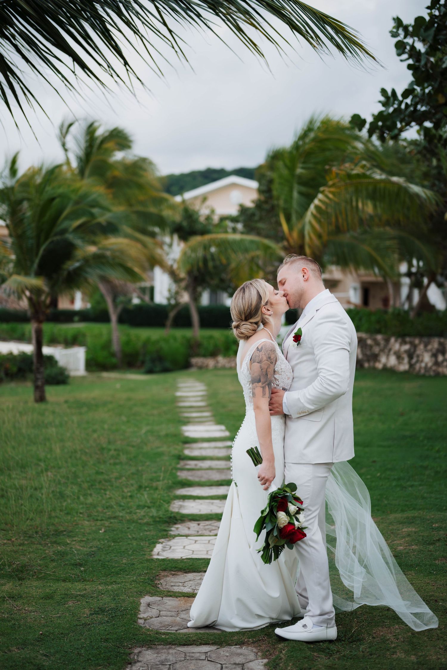 Bride and groom kiss on garden path surrounded by palm trees at tropical destination wedding.