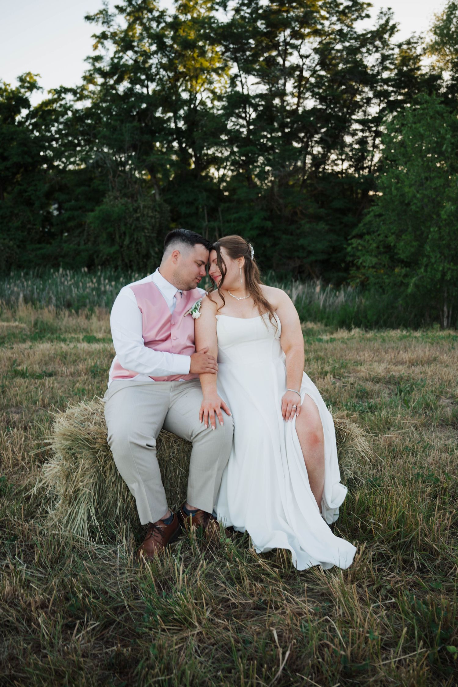 Bride and groom share a romantic moment sitting on a hay bale in a rustic field at golden hour.