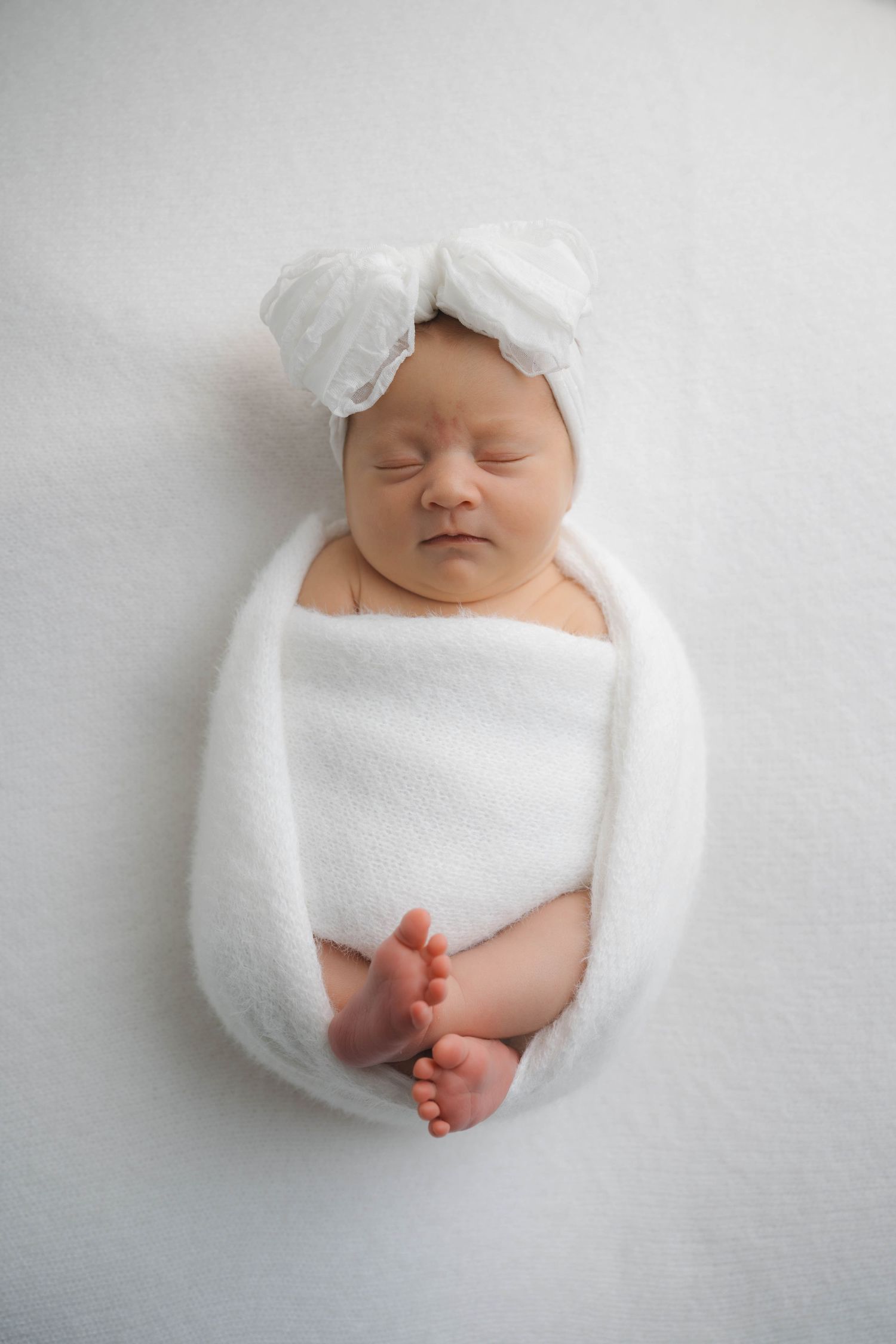 Sleeping newborn baby swaddled in white wrap with bow headband on white background.