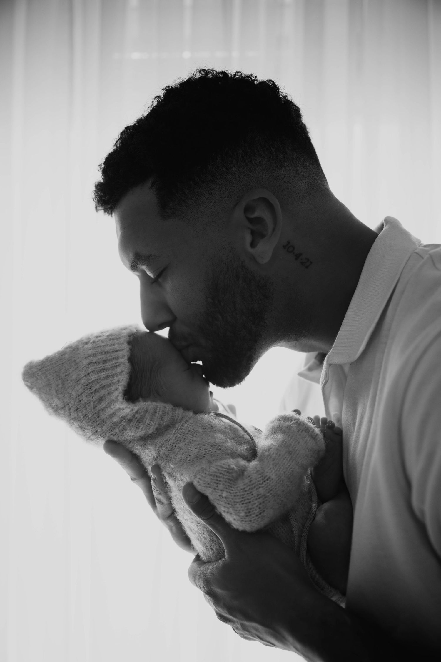 Black and white photo of a father tenderly kissing his newborn baby wrapped in a knit blanket.