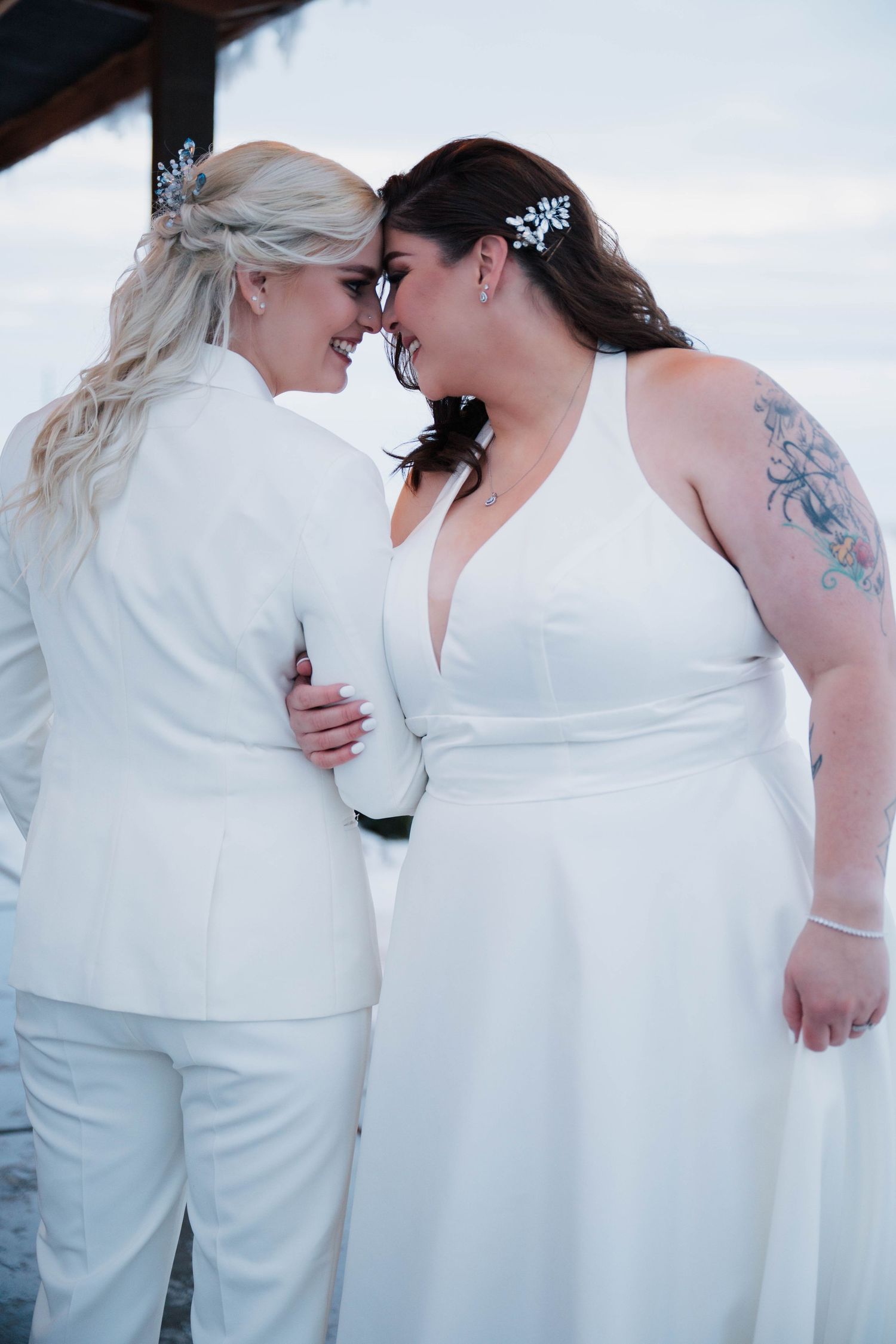 Two brides in white wedding attire share an intimate moment, touching foreheads and smiling at each other outdoors.